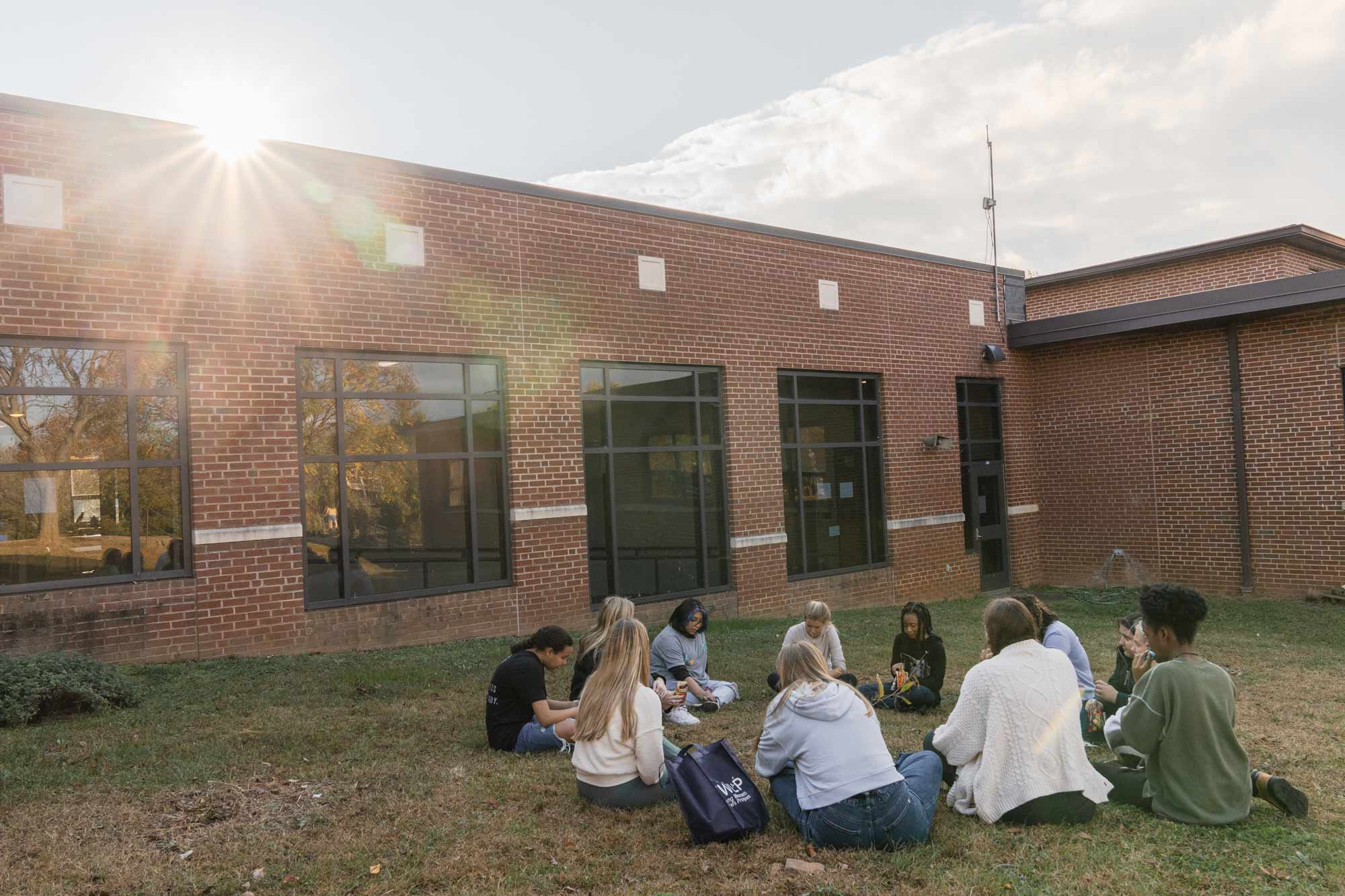 Group of students sitting on the ground in a circle talking outside of a brick school building