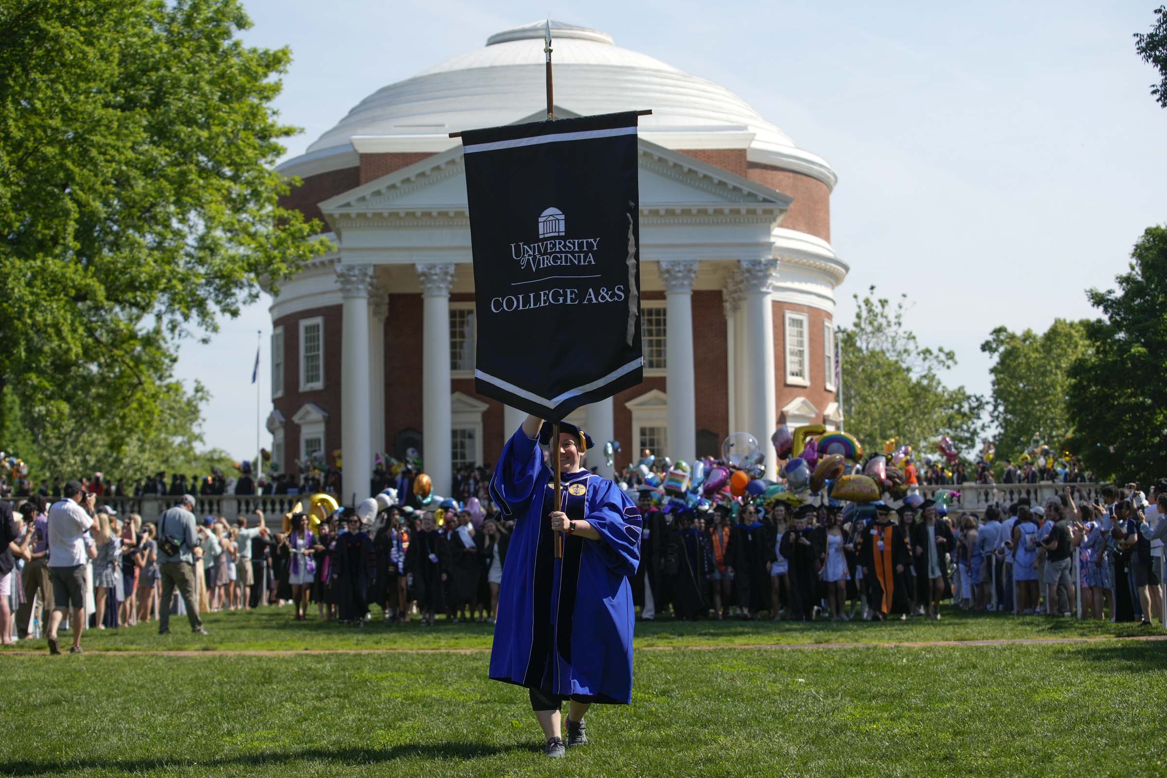 Leading a crowd of graduating students, a woman in a blue robe carries a banner reading University of Virginia College A&S