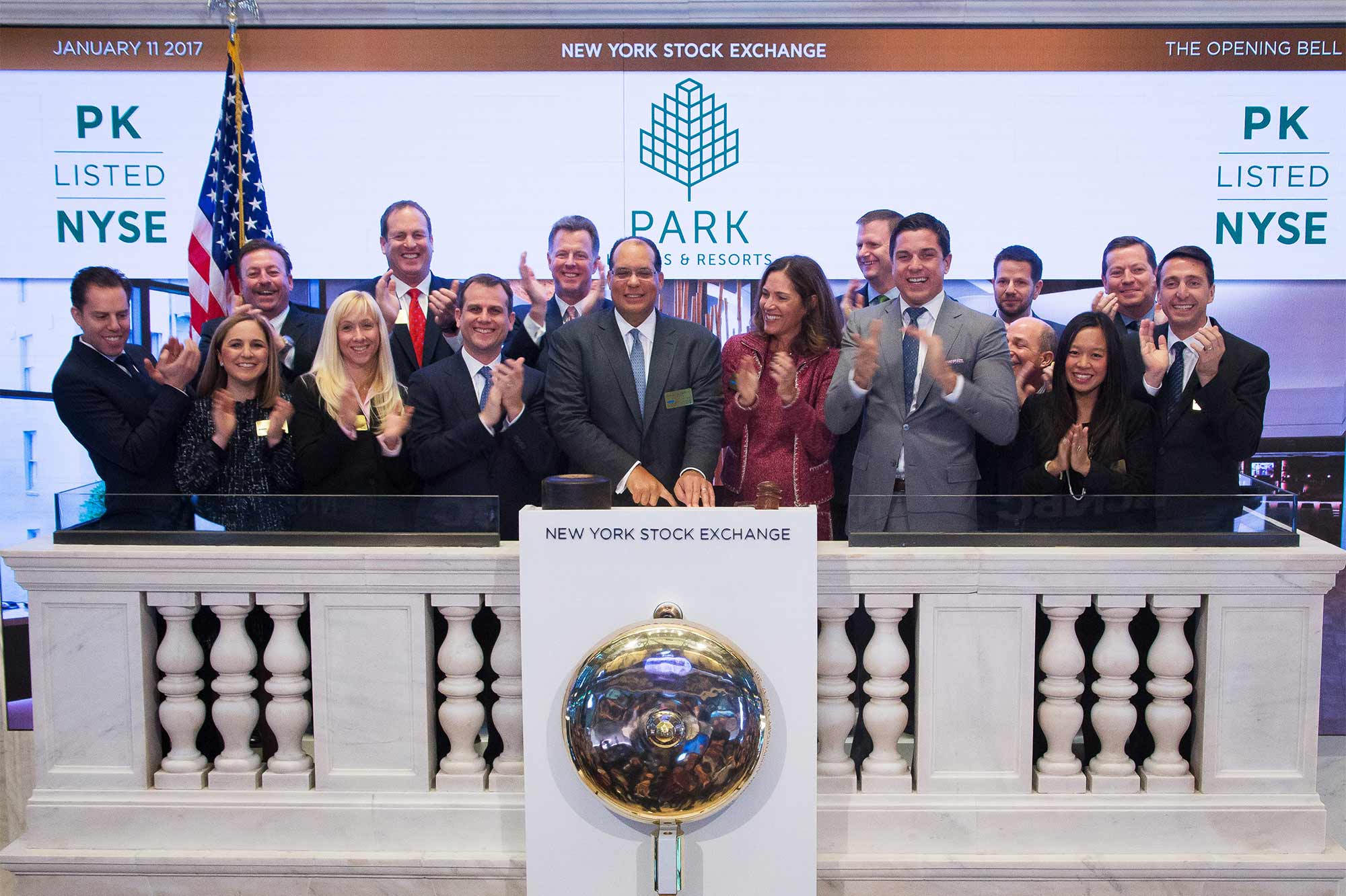 Baltimore stands behind a podium labeled New York Stock Exchange, with 15 other people, clapping and smiling