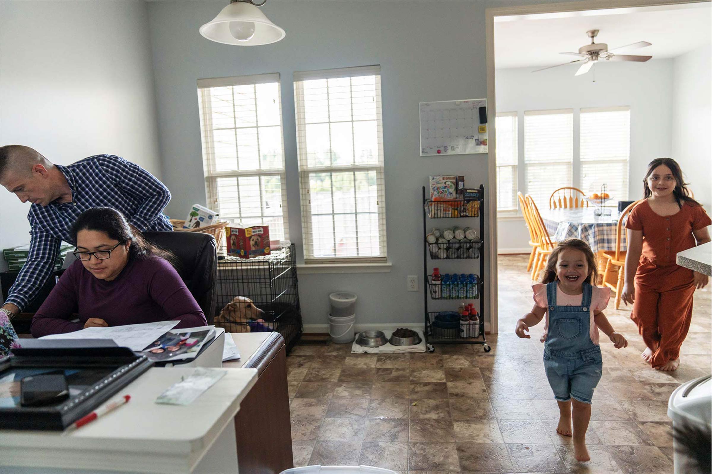 A woman studies at a desk while a young girl chases her little sister across the living room