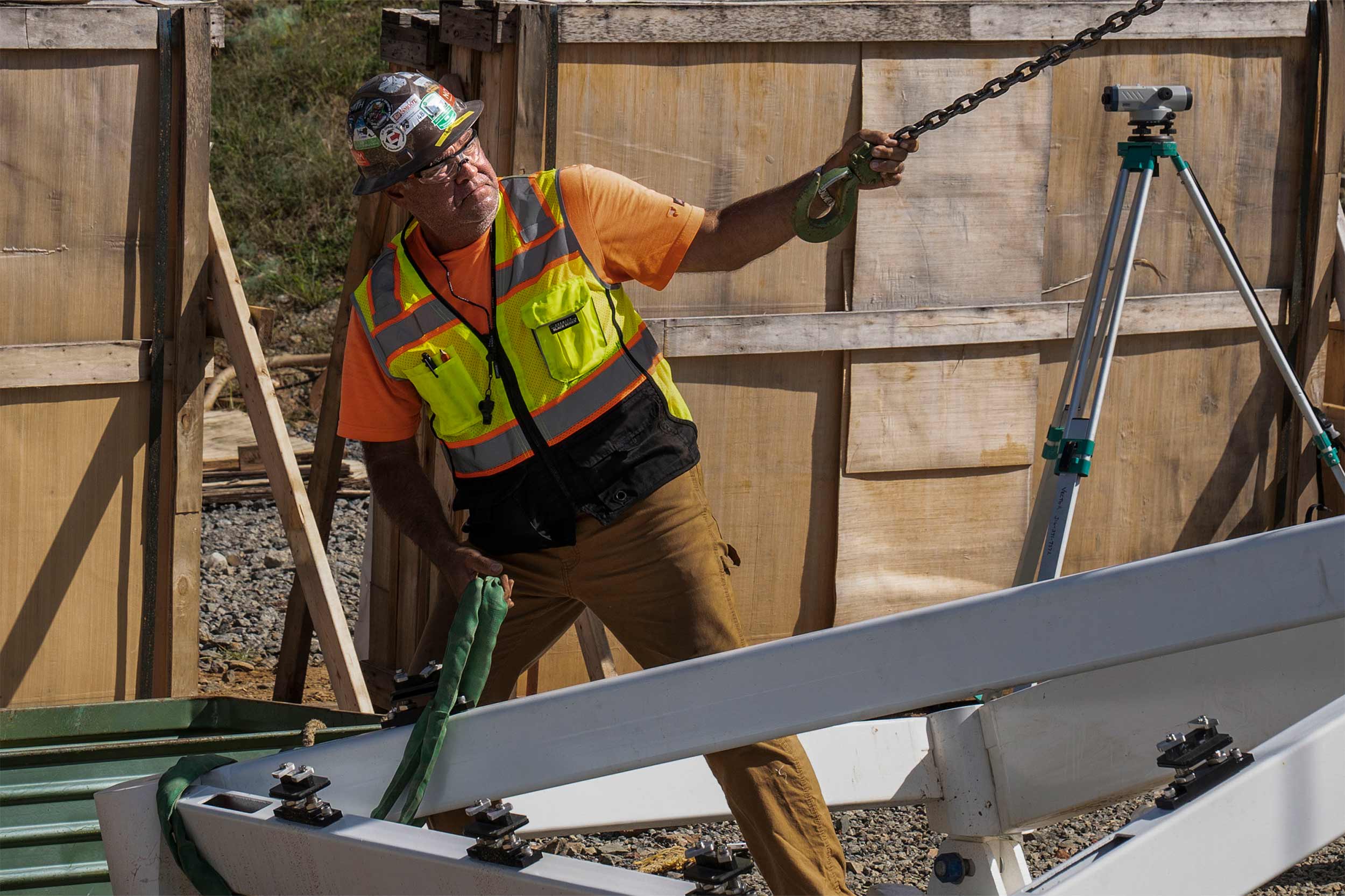 A construction worker in a safety vest and hard hat holds a large hook on a chain in one hand and a strap secured around the corner of a frame in the other