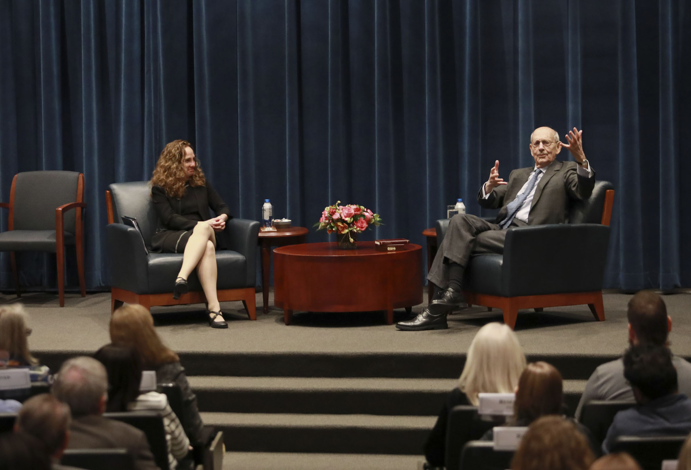 Breyer talks and gestures while seated in a leather chair on a stage. Goluboff listens from another chair.
