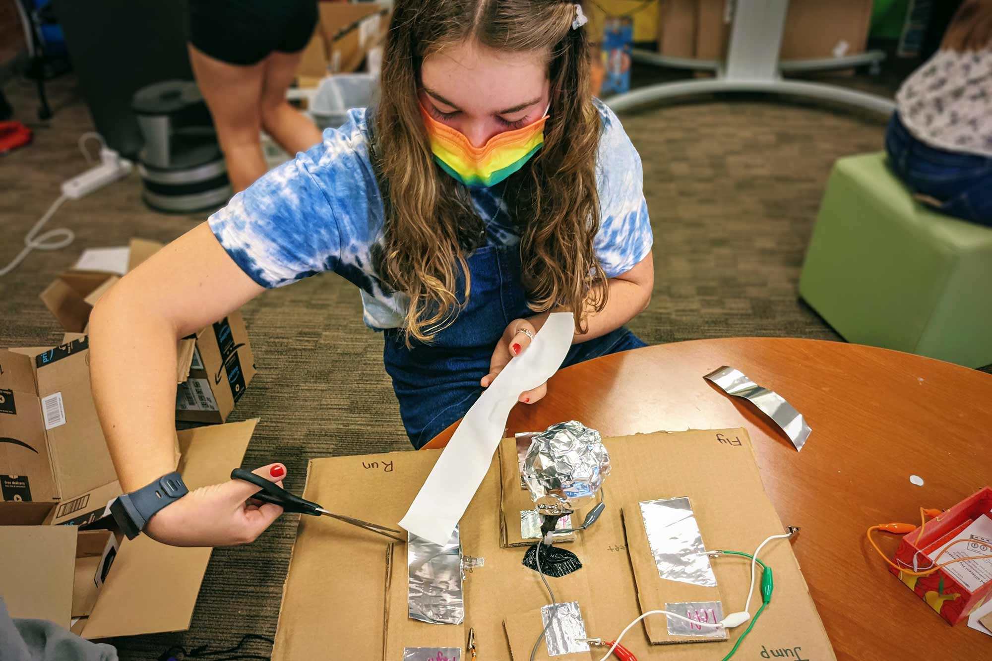 A girl in a rainbow mask cuts a strip of aluminum foil attached to a cardboard contraption