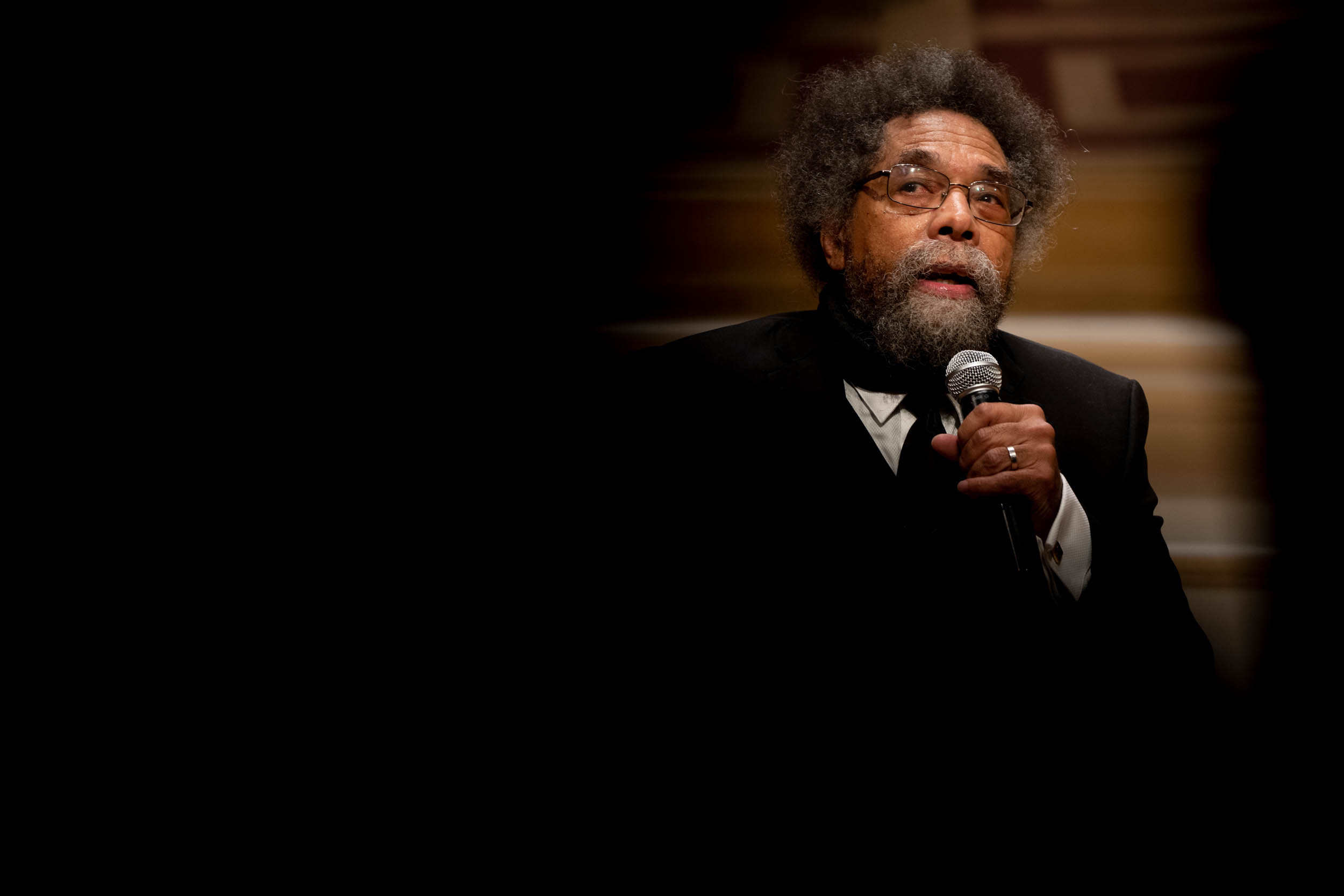 Cornel West speaking into a microphone with most of the image dark except for him