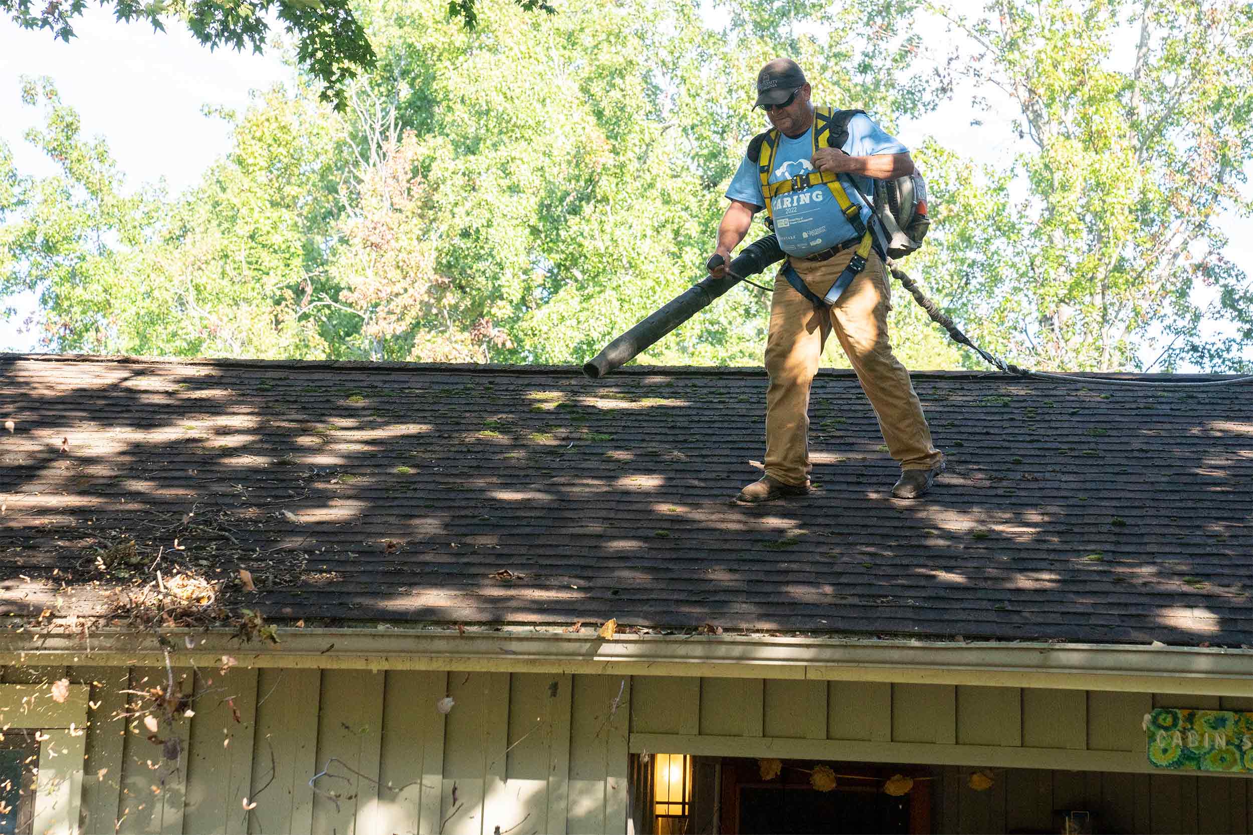 A man with a leaf blower on a building roof cleans out the gutters.