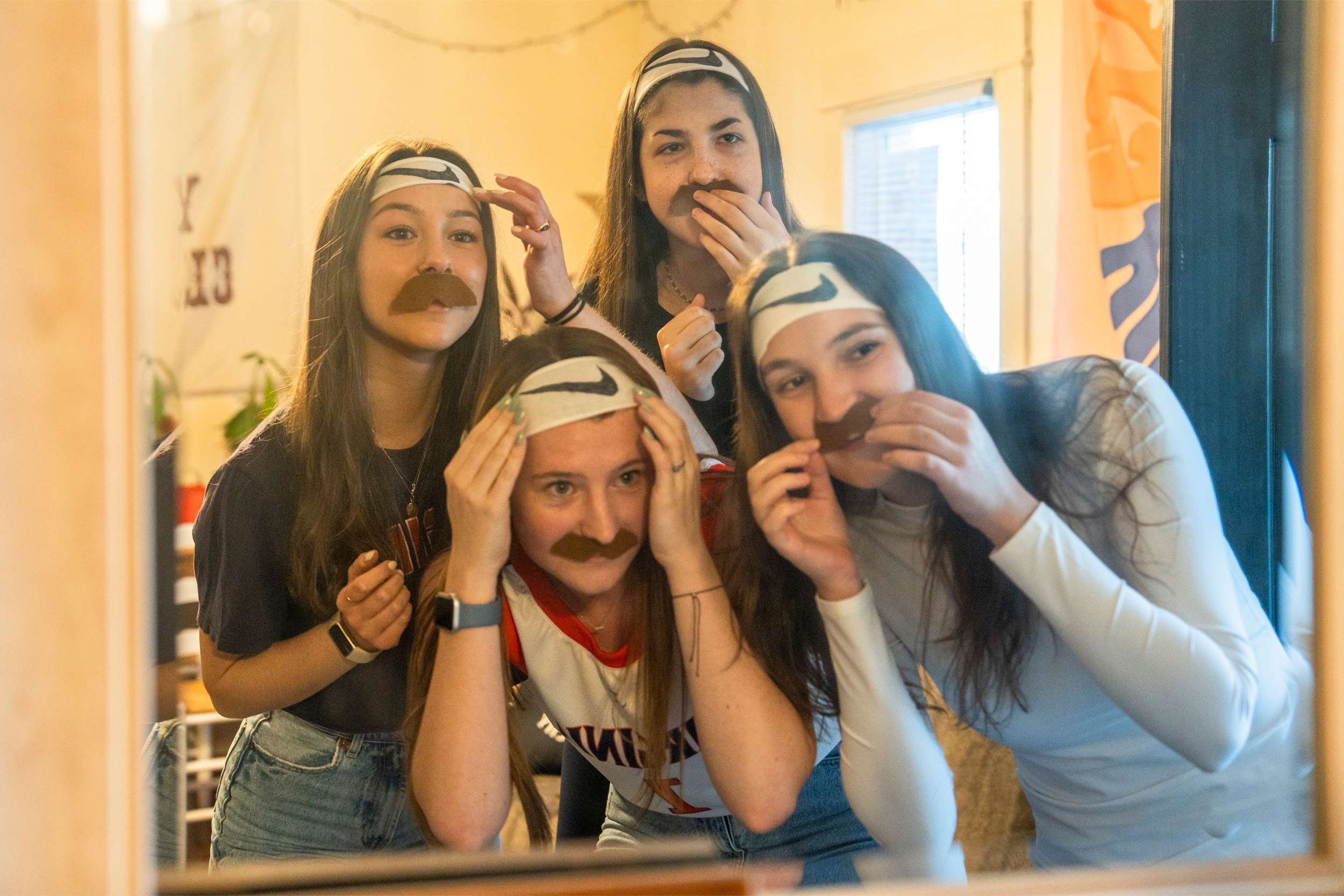 Four female UVA students looking in the mirror as they put on their headbands and fake mustaches