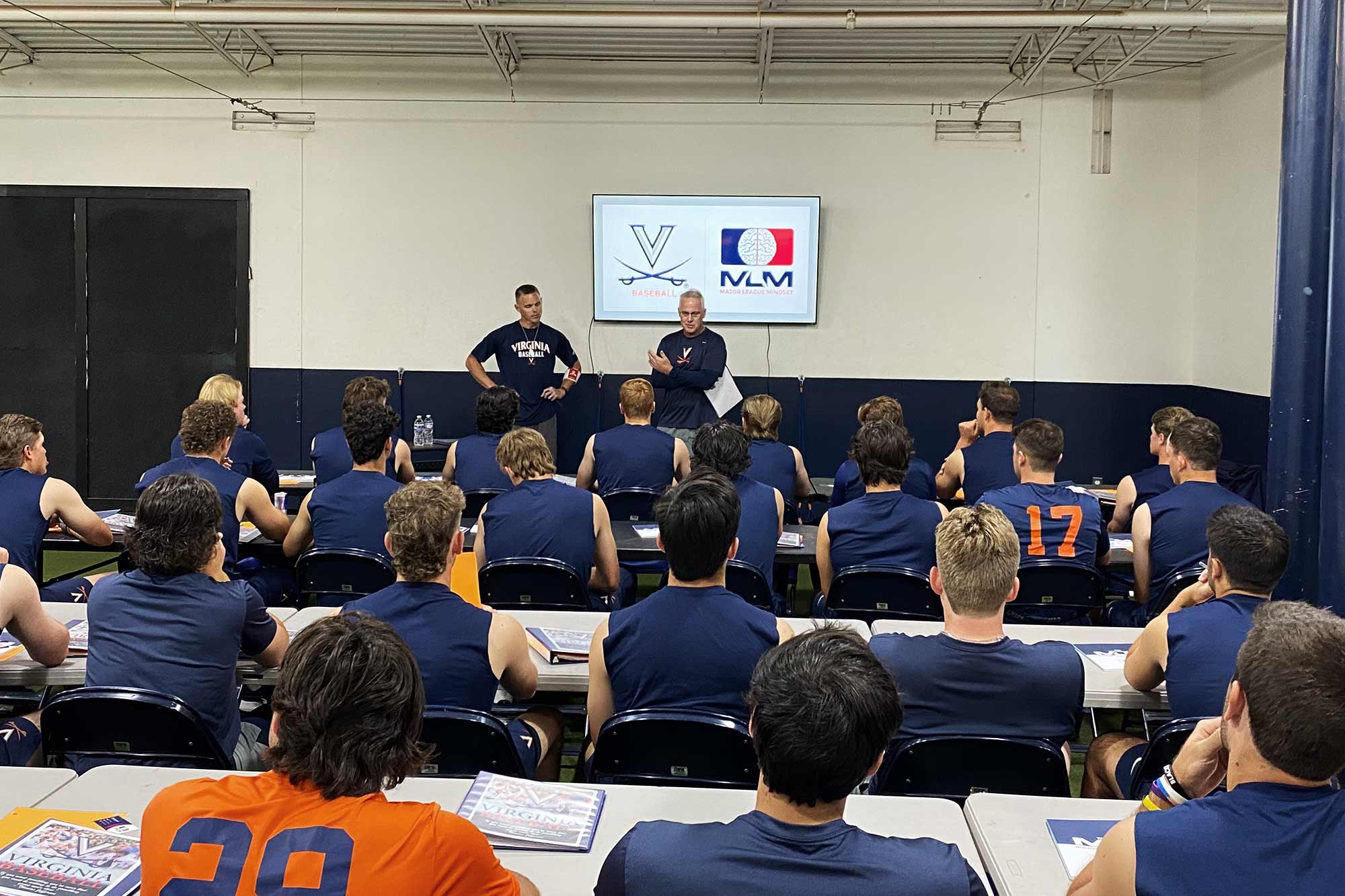 Two coaches at the front of a room filled with rows of seated players