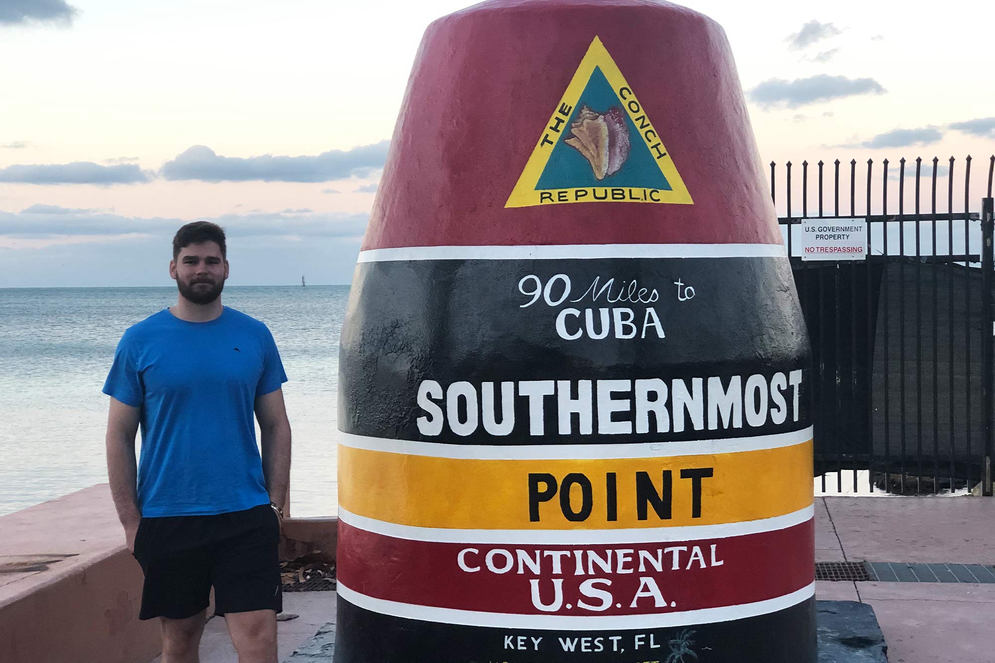 Pleško stands next to a red, black, and yellow monument in front of the ocean that reads "90 miles to Cuba Southernmost Point Continental U.S.A. Key West, FL"