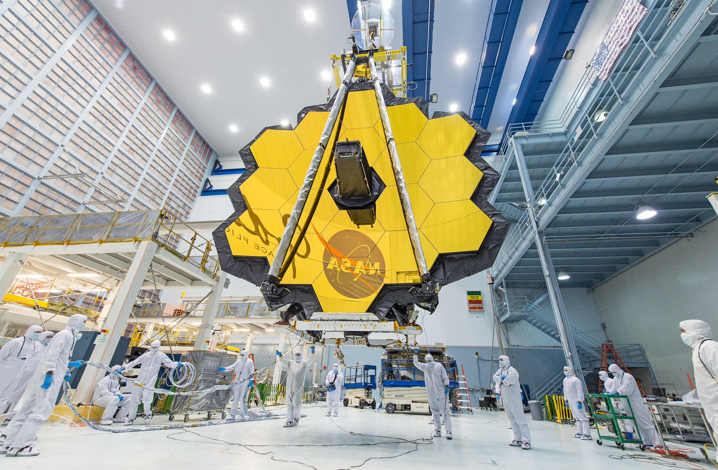 People in clean suits are dwarfed by the massive Webb Telescope and its 18 golden mirrors.