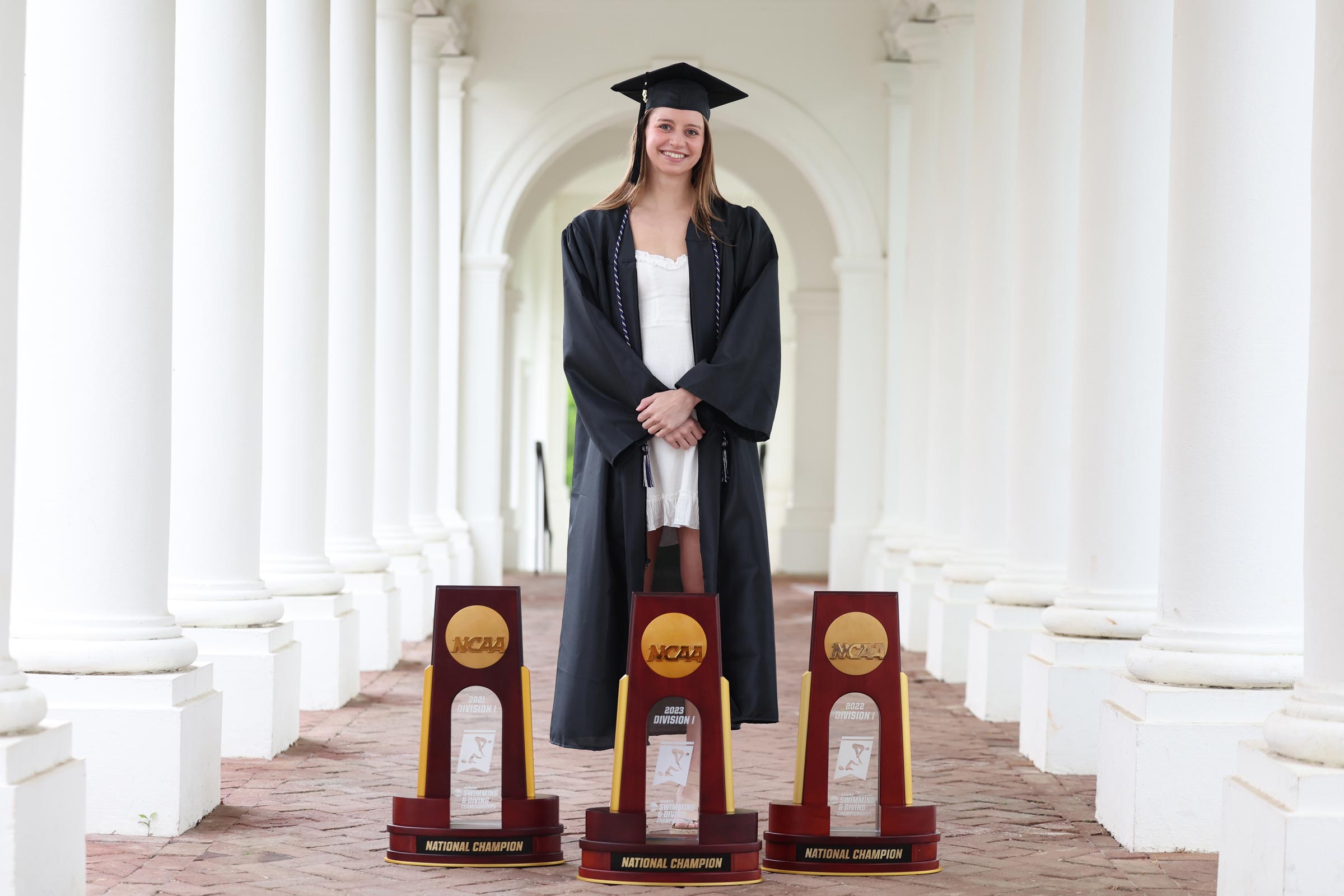 Kate Douglass in her UVA cap and gown with three trophies in front of her