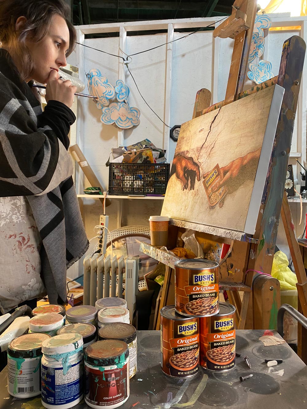 A woman holding a paint brush regards a painting on an easel, a parody of Michelangelo's 'The Creation of Adam,' in which God's hand is holding a can of Bush's baked beans