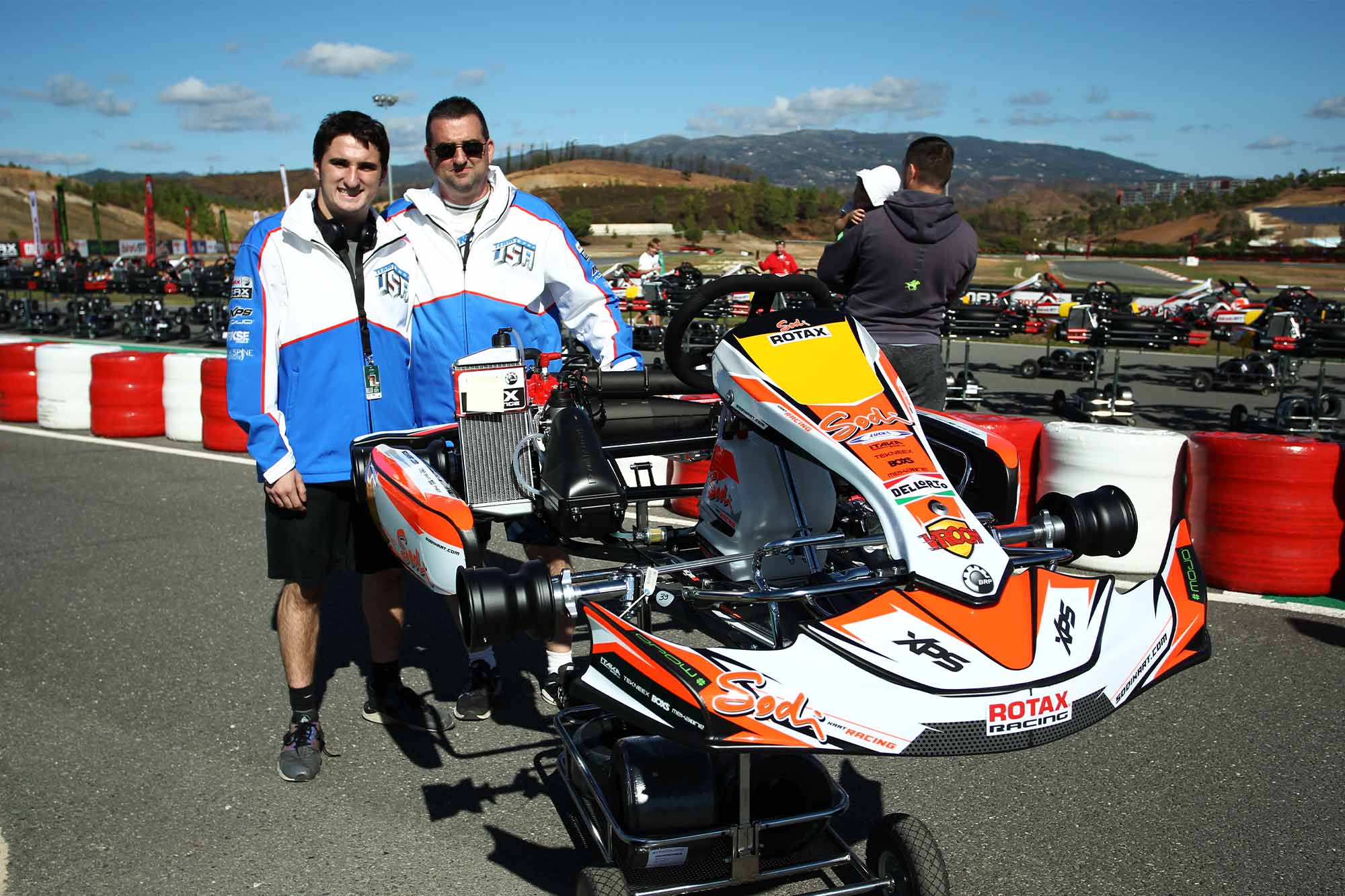 Two men stand in behind a go-kart at a race track