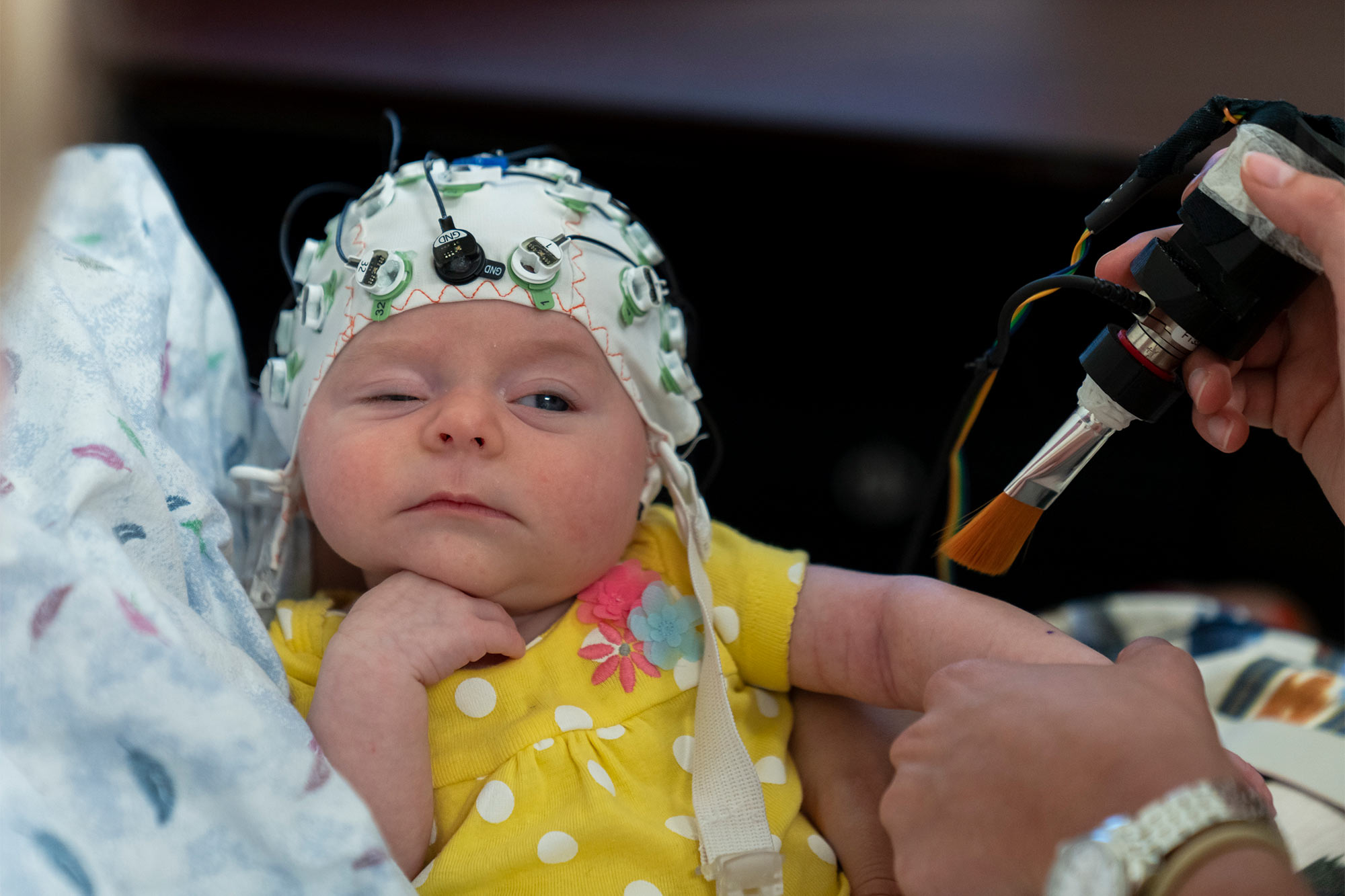 A baby wearing a skullcap rests her chin on her hand, while looking with one eye open at a researcher holding a paintbrush near her arm