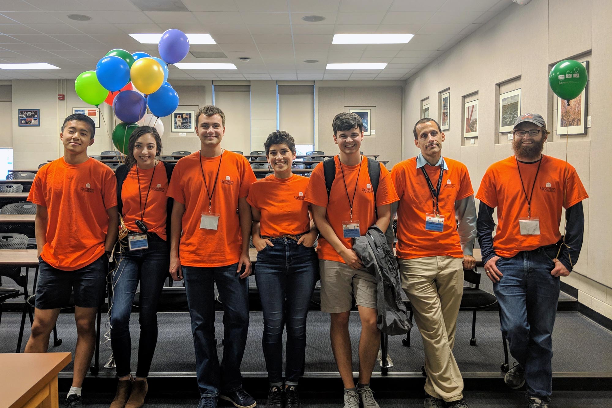 Group photo of seven people wearing orange shirts and name-tags in a large conference room