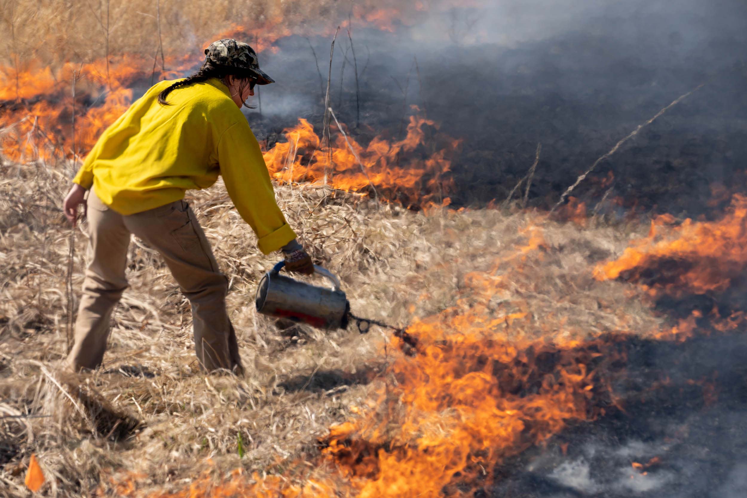 A woman pours burning fuel at the fiery edge of a field of burnt thatch