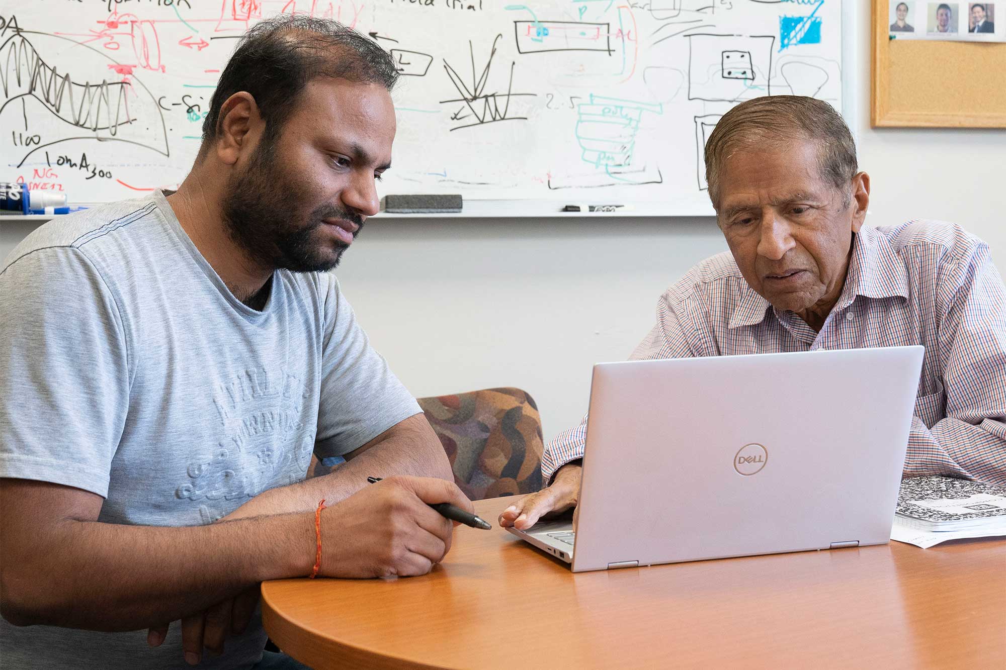 In front of a whiteboard covered in hand-drawn schematics, Kanaujia and Gupta sit at a desk looking at a laptop together