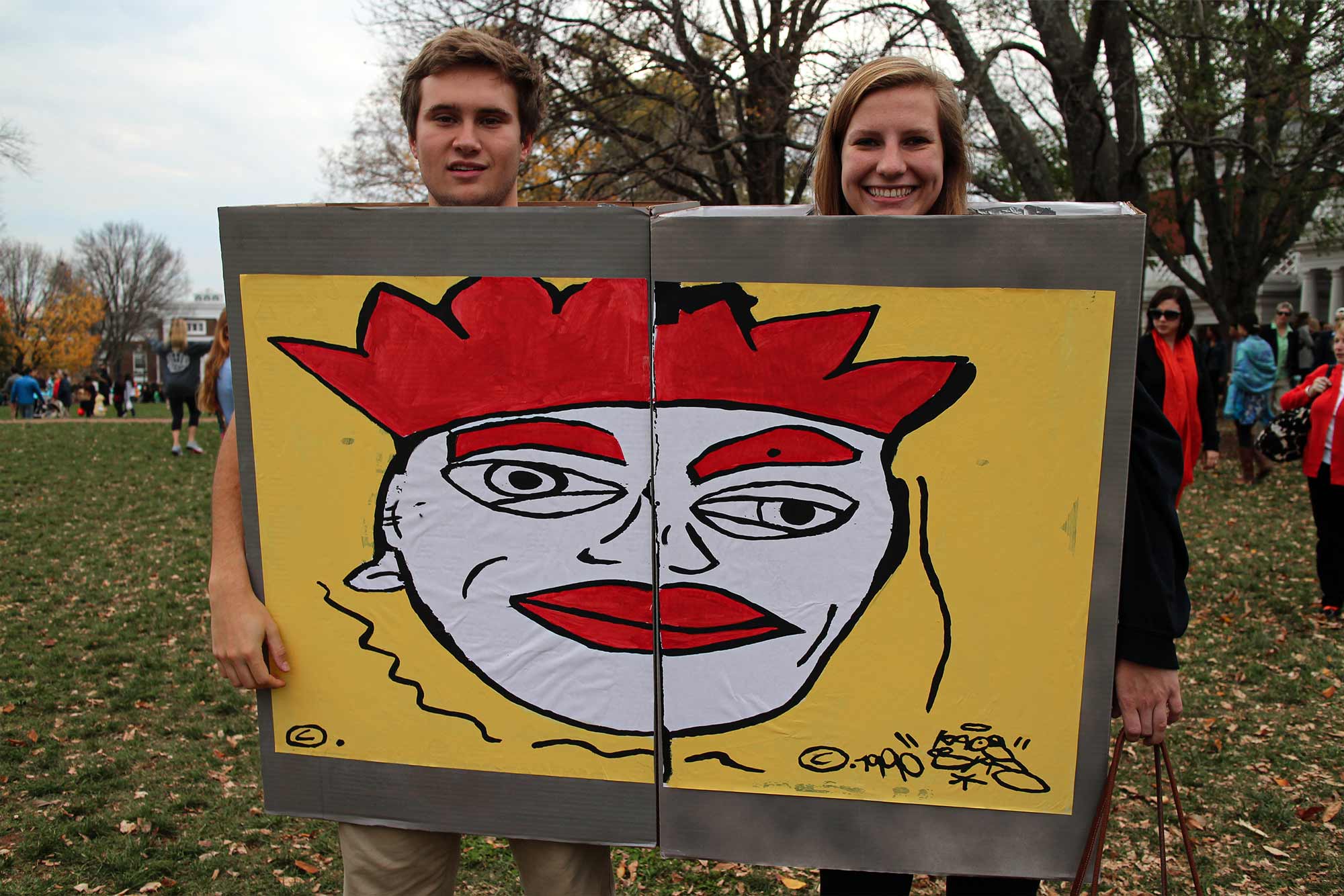 Two students in Halloween costumes that together make a piece of a section of the Berlin wall on Grounds, a white face with rest lips, eyebrows, and crwon on a yellow background