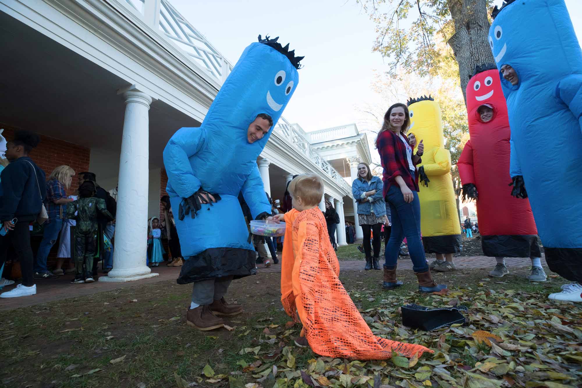 A student in a blue tube man costume gives candy to a child in an orange cape