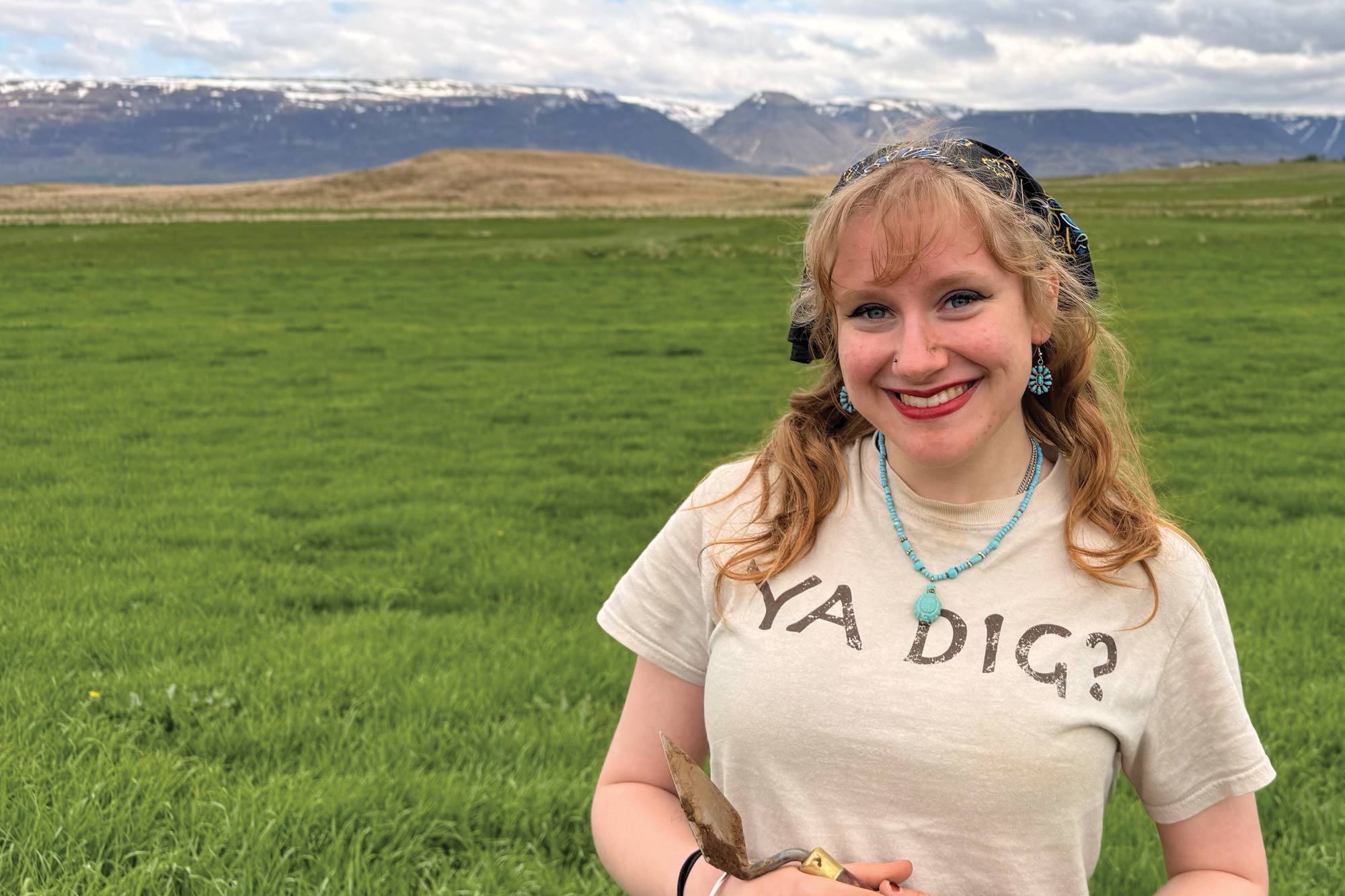 Latham holding a trowel in a green field with mountains in the background.