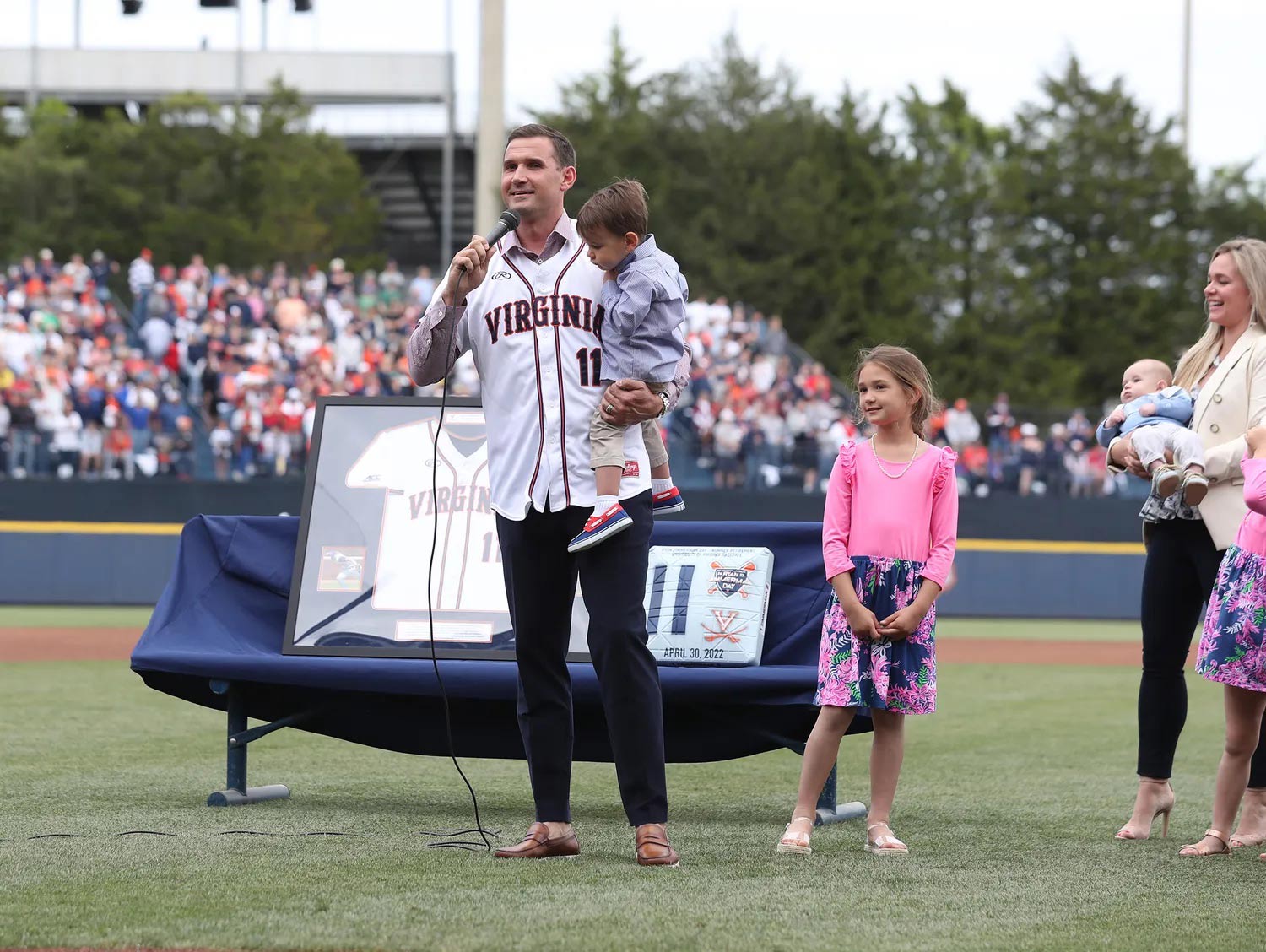 Ryan Zimmerman in a Virginia jersey stands in a baseball field holding his small son and speaking into a microphone