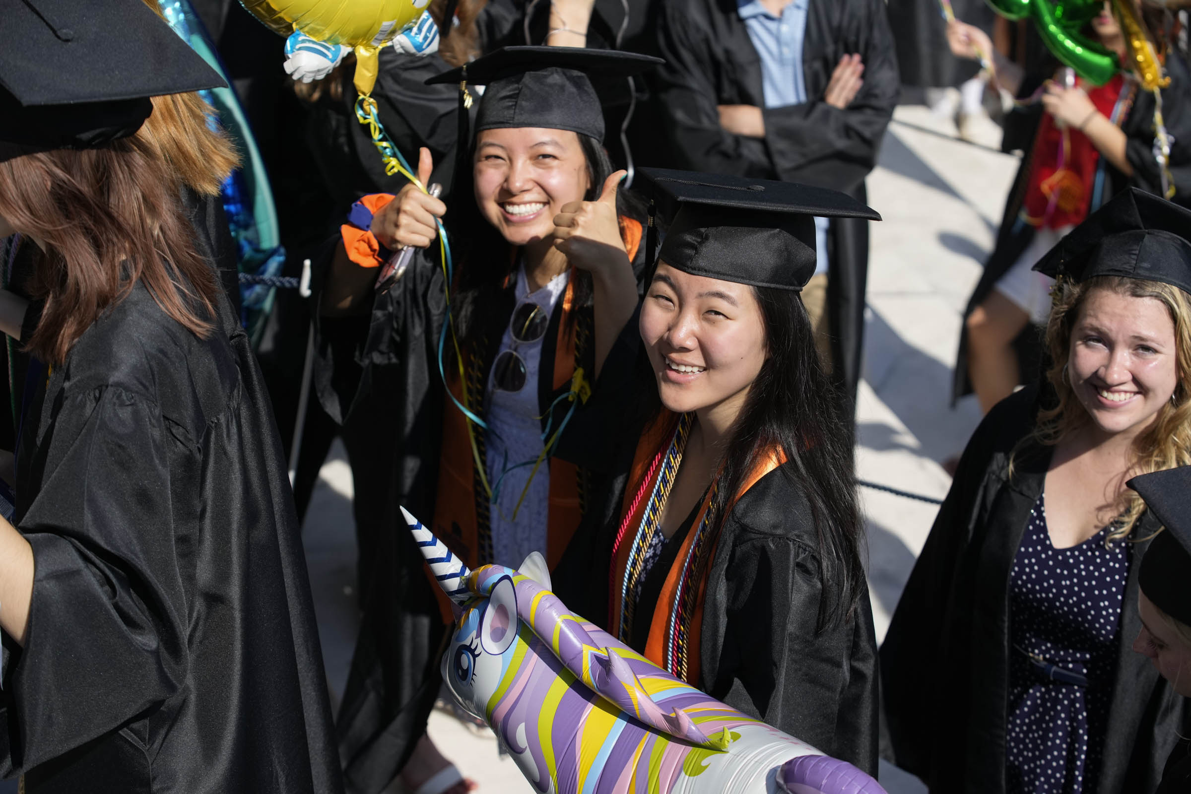 A graduating student gives two thumbs up, with two other students looking on.