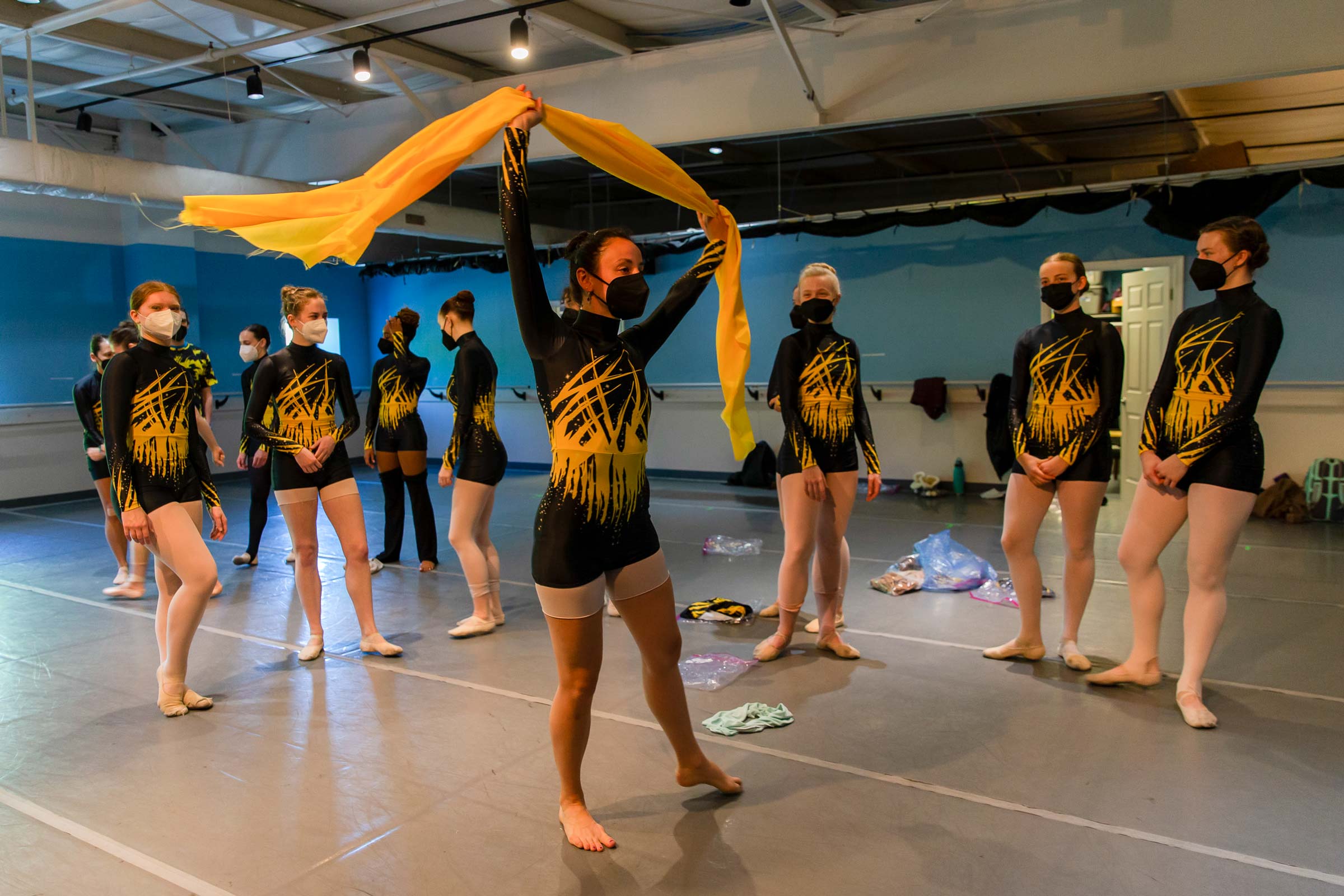 Ballet dances in black and yellow body suits stand in a half-circle, watching a woman dance while holding a yellow scarf