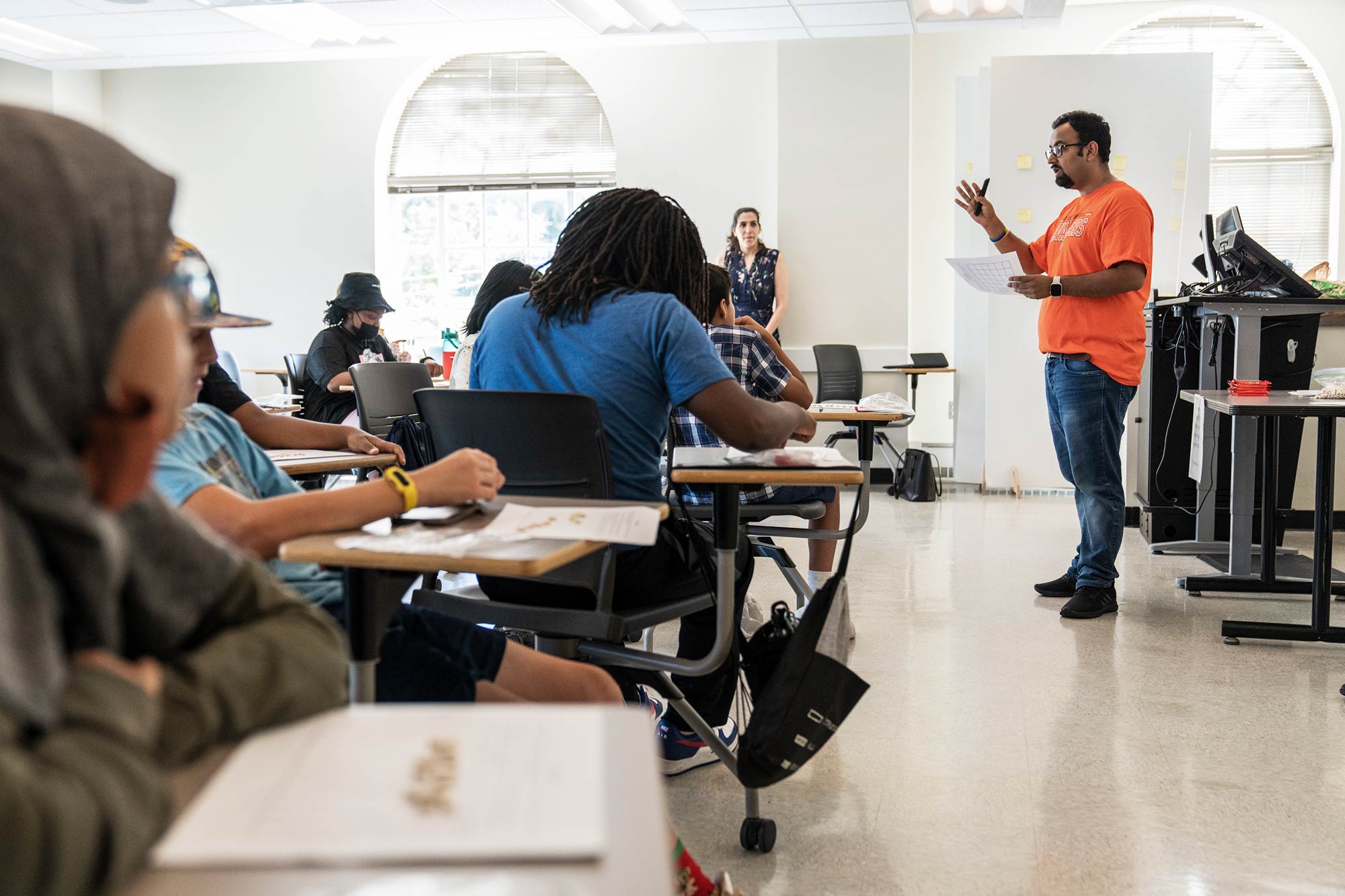 A man in an orange shirt talks to students seated at desks