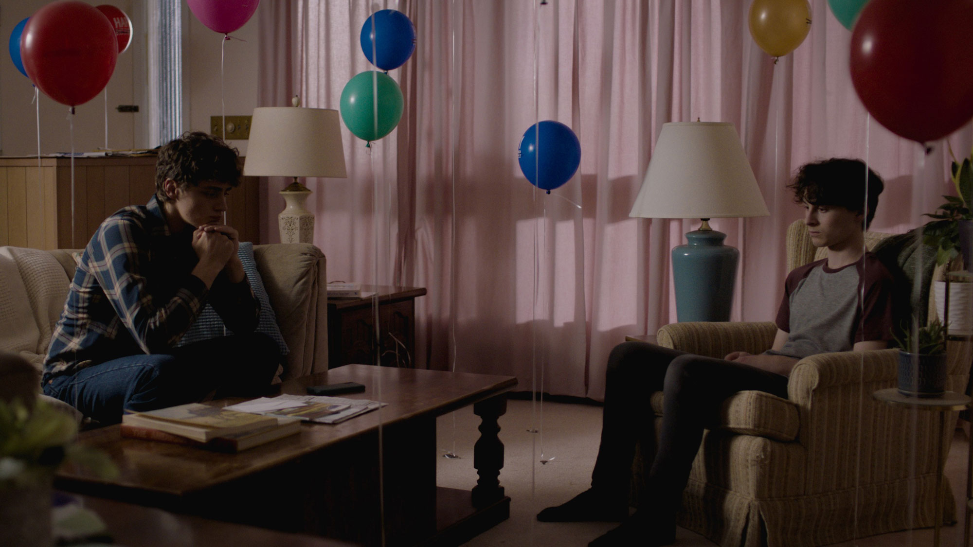 Two somber young people sit in a living room filled with colorful balloons