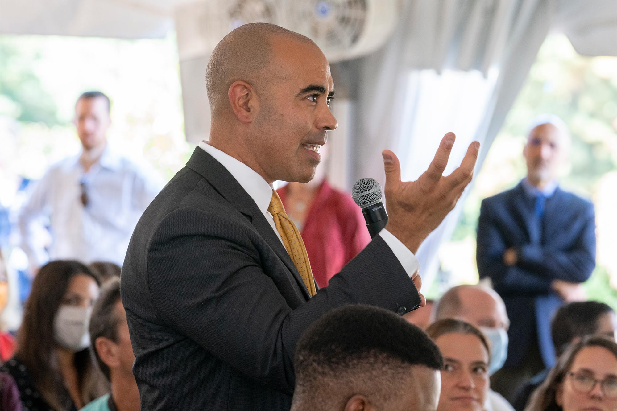 Ian Solomon speaks into a microphone from the middle of a seated crowd under a tent