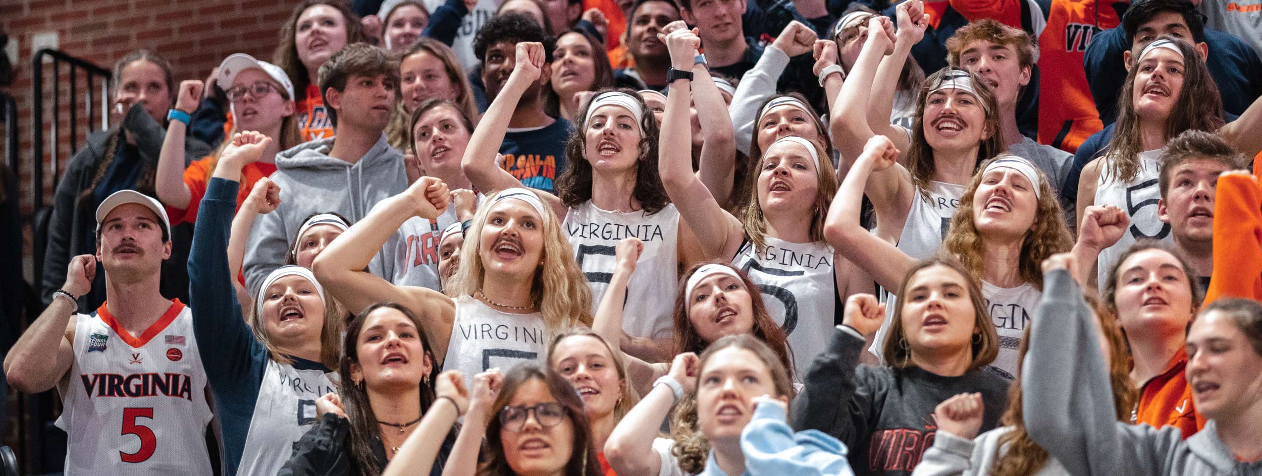 A crowd of students wearing headbands, fake mustaches, and sleeveless white tee shirts with the number 5 written on them cheering at a UVA Men's Basketball game
