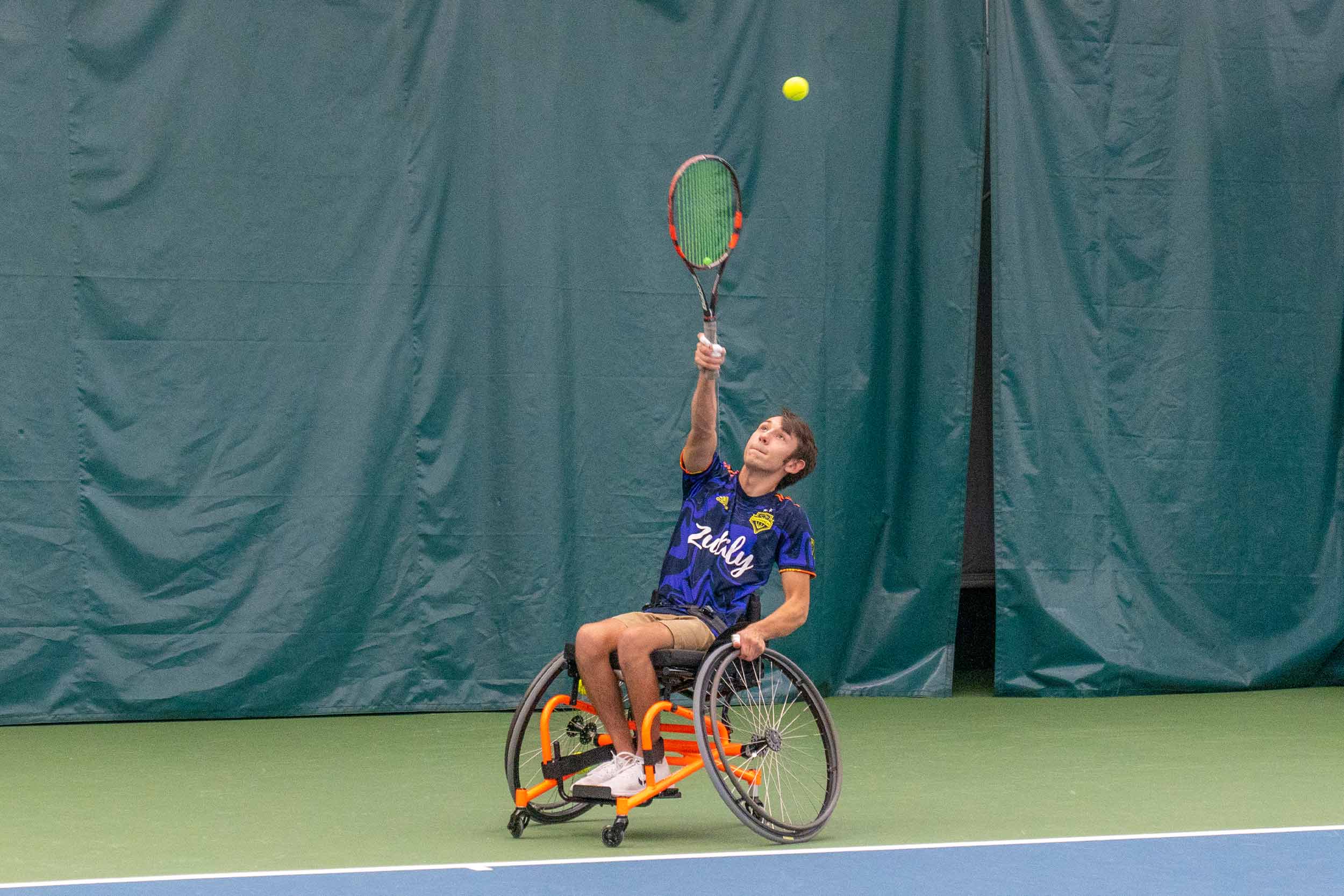 Man in wheelchair serves tennis ball.