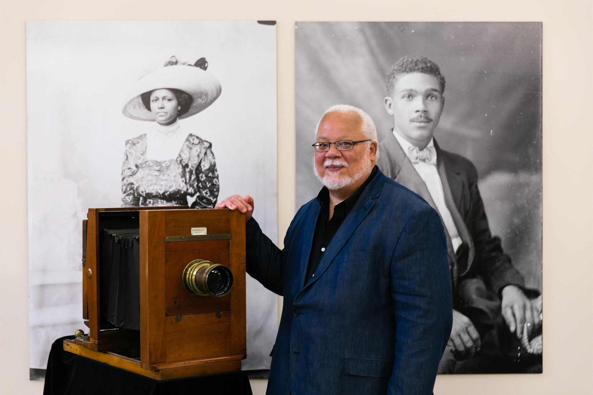 John Edwin Mason stands in front of two large portraits, resting a hand on an antique camera
