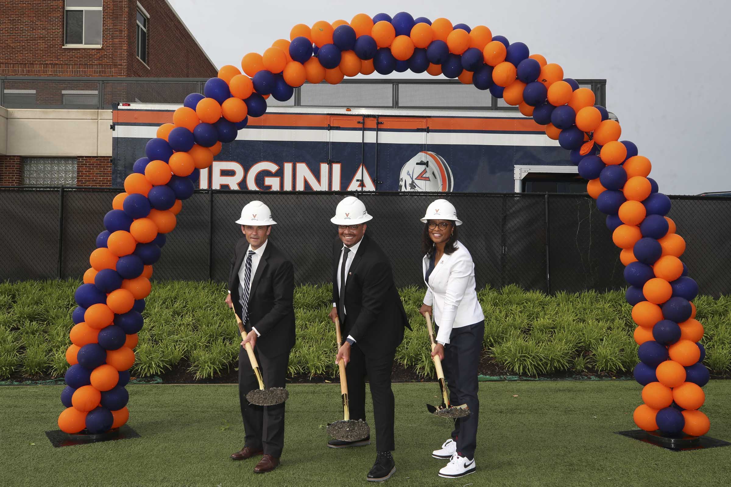 Three people in hard hats pose with shovels under a blue and orange balloon arch