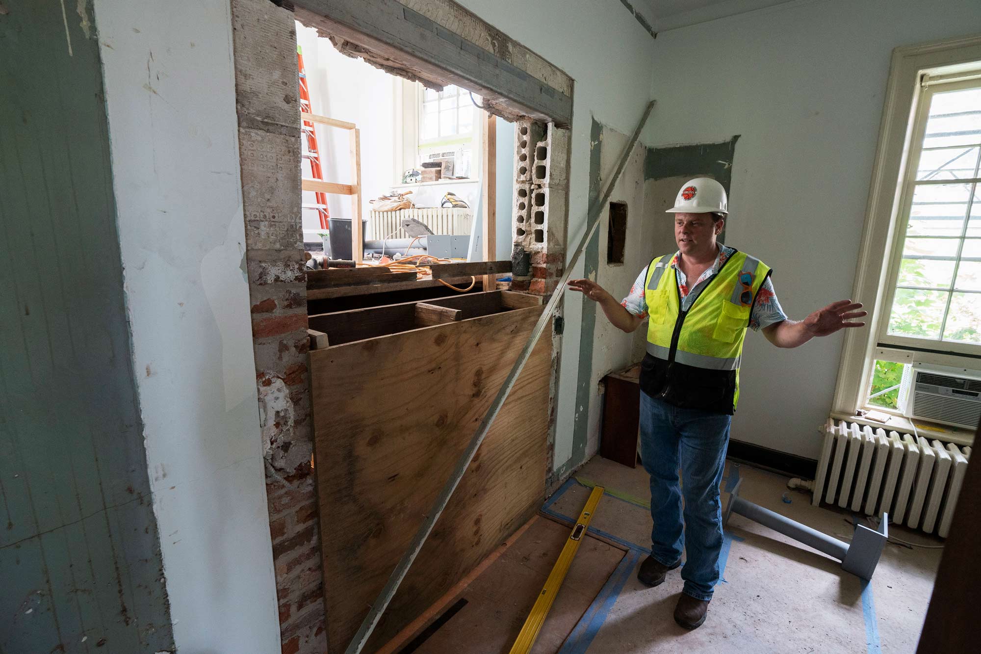 James Zehmer stands in the lower Lawn Room 48, wearing a high visibility vest and hard hat, next to a hole in the wall revealing Room 50.