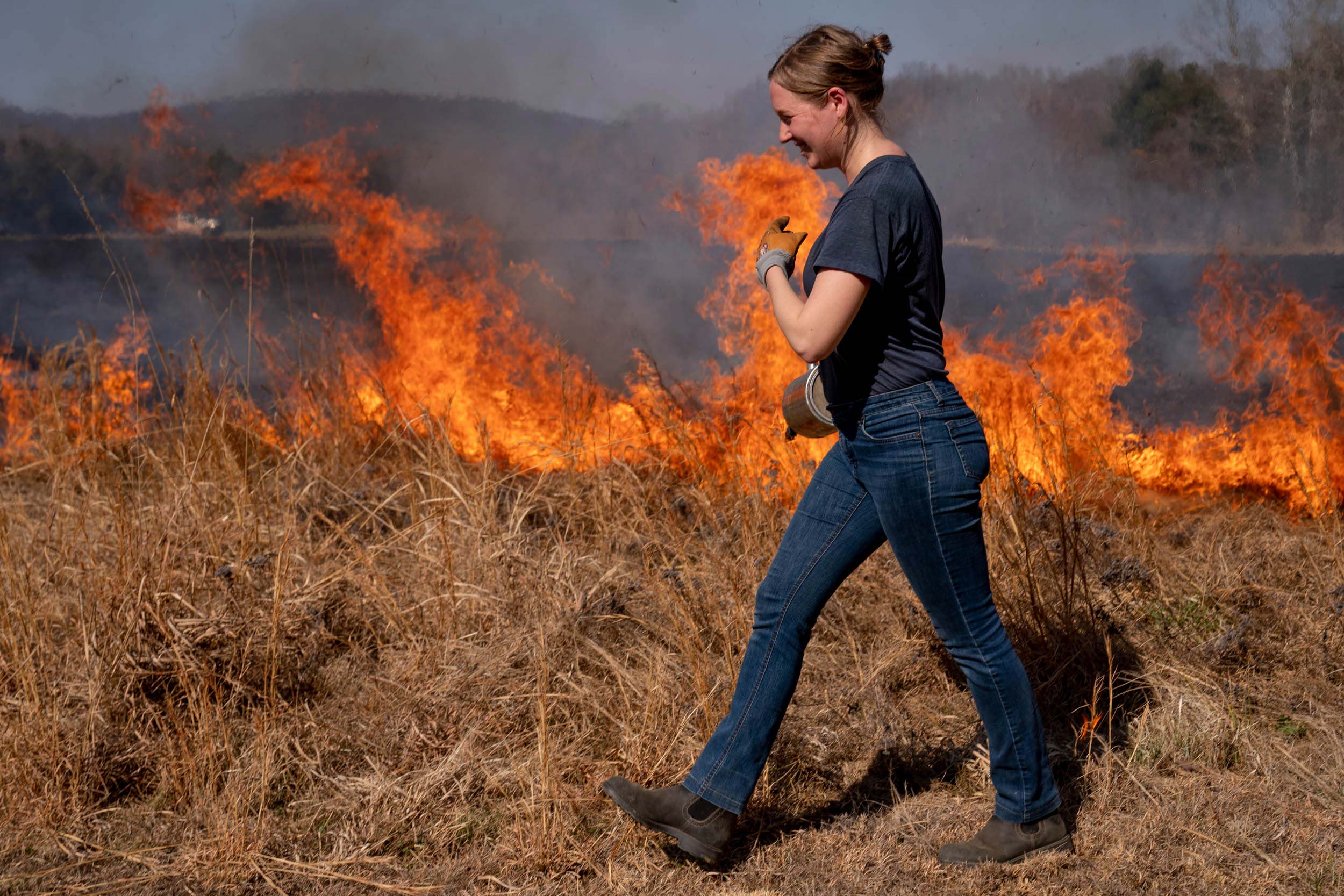A woman walks next to a line burning thatch in front of a lake