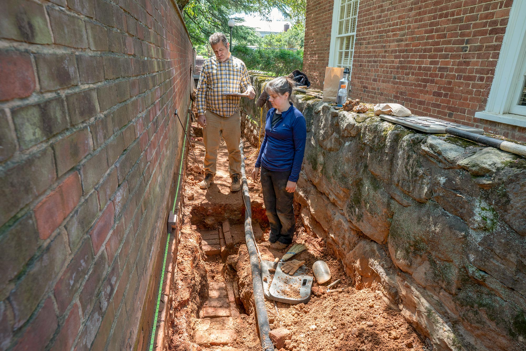 A man and a woman in work clothes stand in the dug-up path and examine bricks in the ground