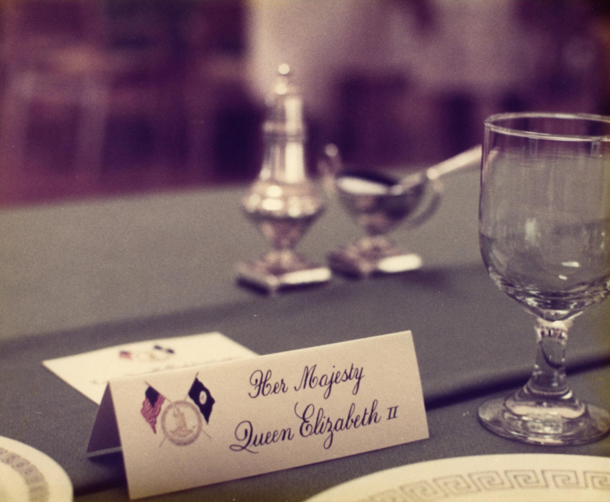 A placard reading Her Majesty Queen Elizabeth II sits on a table beside a glass goblet and silver serve-ware