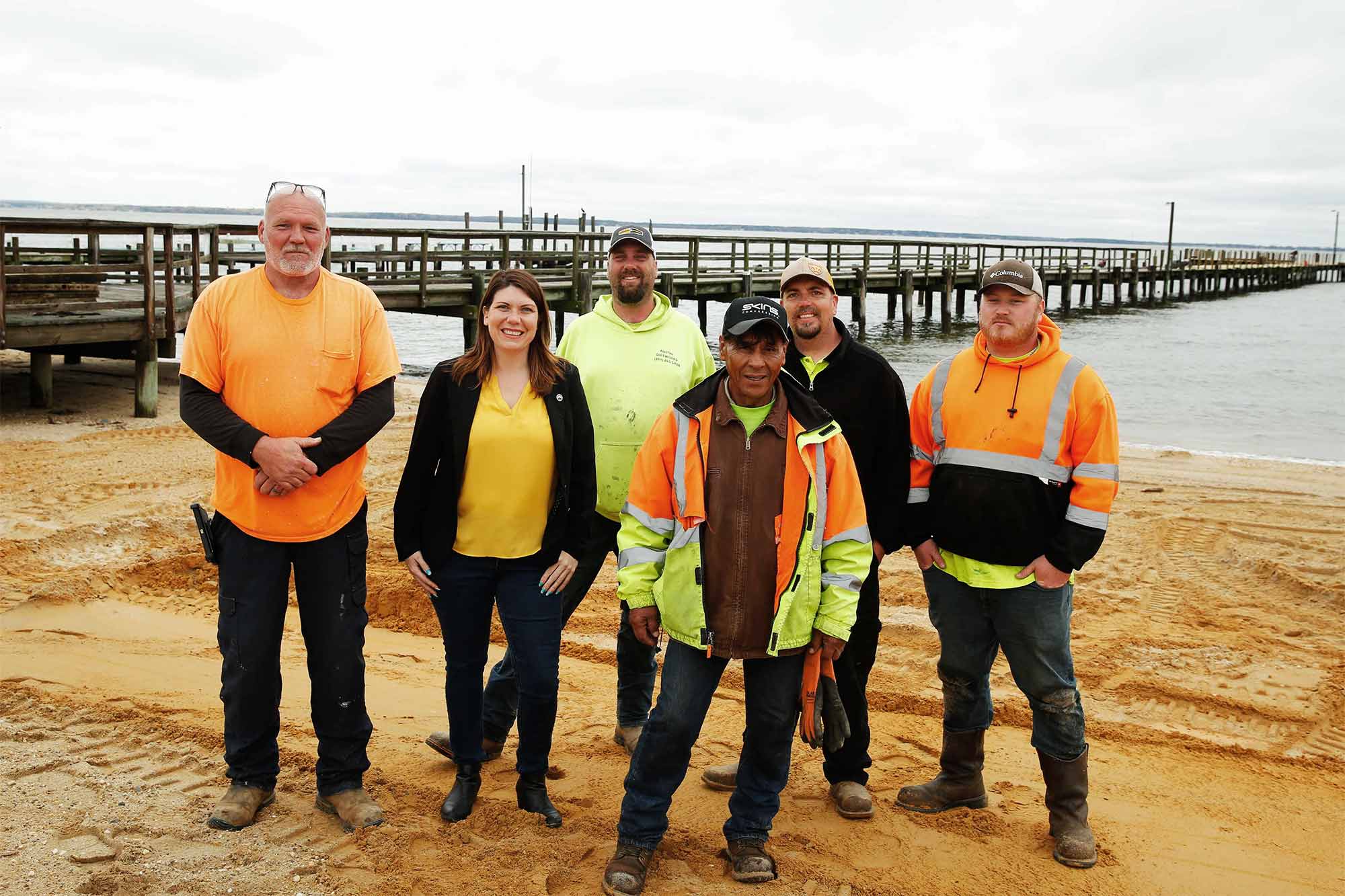 A group of people in safety colors poses for a portrait in the sand in front of a long pier
