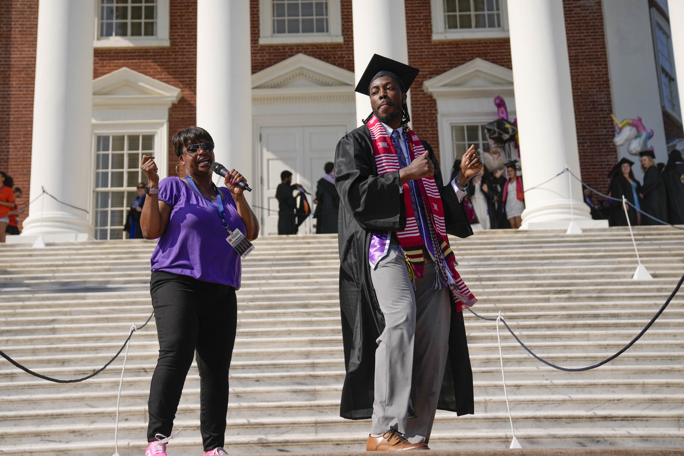 On the steps of the Rotunda, a woman sings into a microphone, while a graduating student dances along