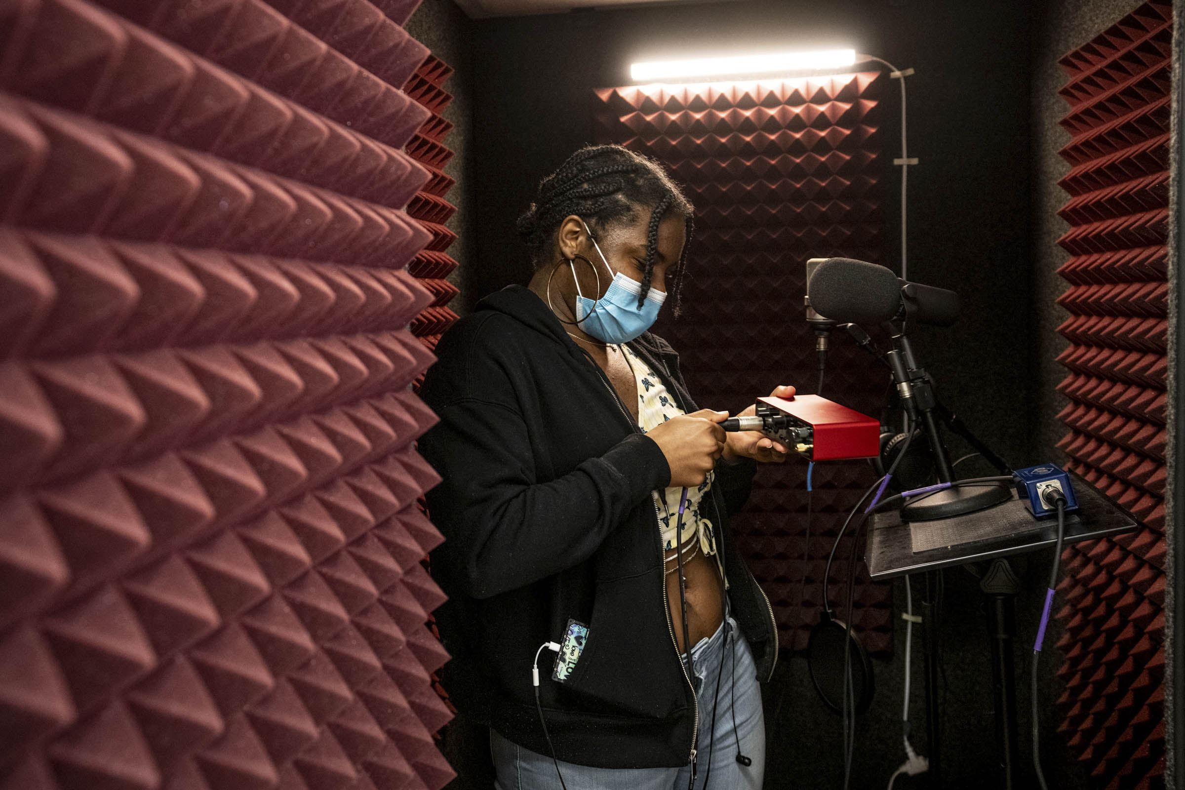 Inside a vocal booth lined with red acoustic foam, a young woman plugs in a microphone cable