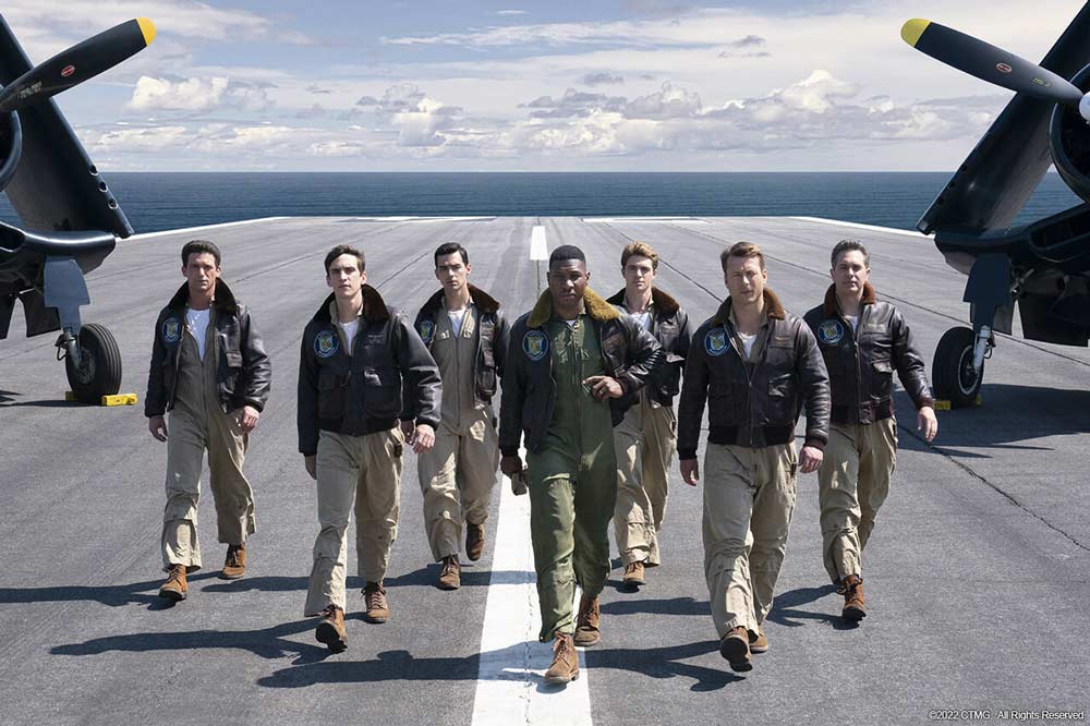 Seven men in bomber jackets walk down an airstrip toward the camera