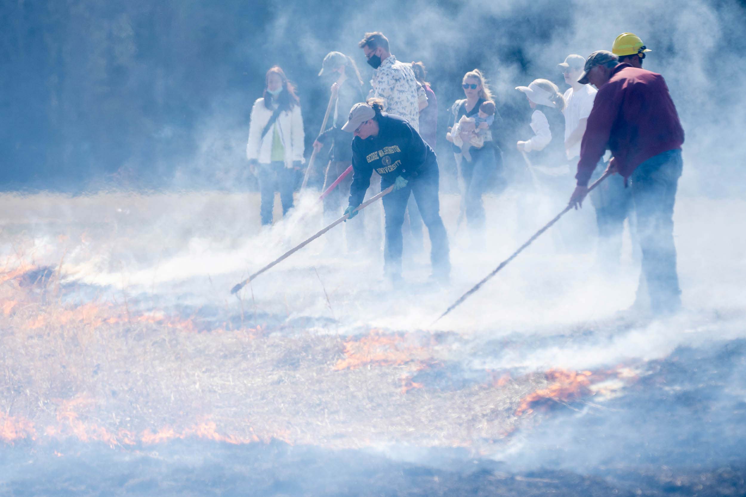 Two people with hoses spray the edge of a fire, while a crowd of people look on through a smoky haze