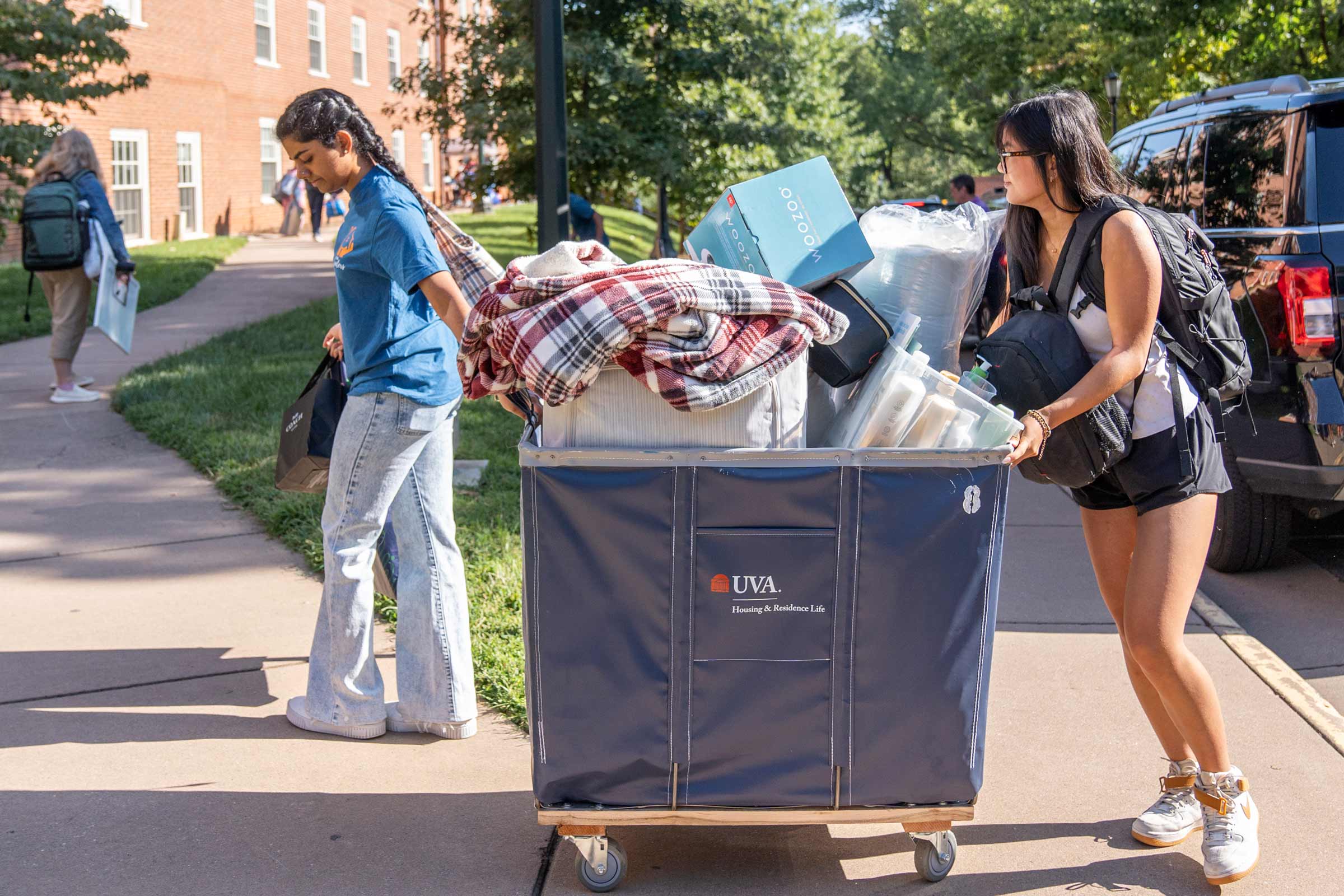 Two students wheel a cart of belongings away from their car toward the dorms