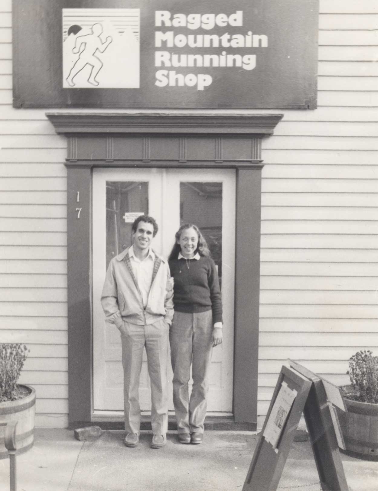 Black and white photo of Mark and Cynthia Lorenzoni standing in front of the entrance to a shop with the sign 'Ragged Mountain Running Shop'