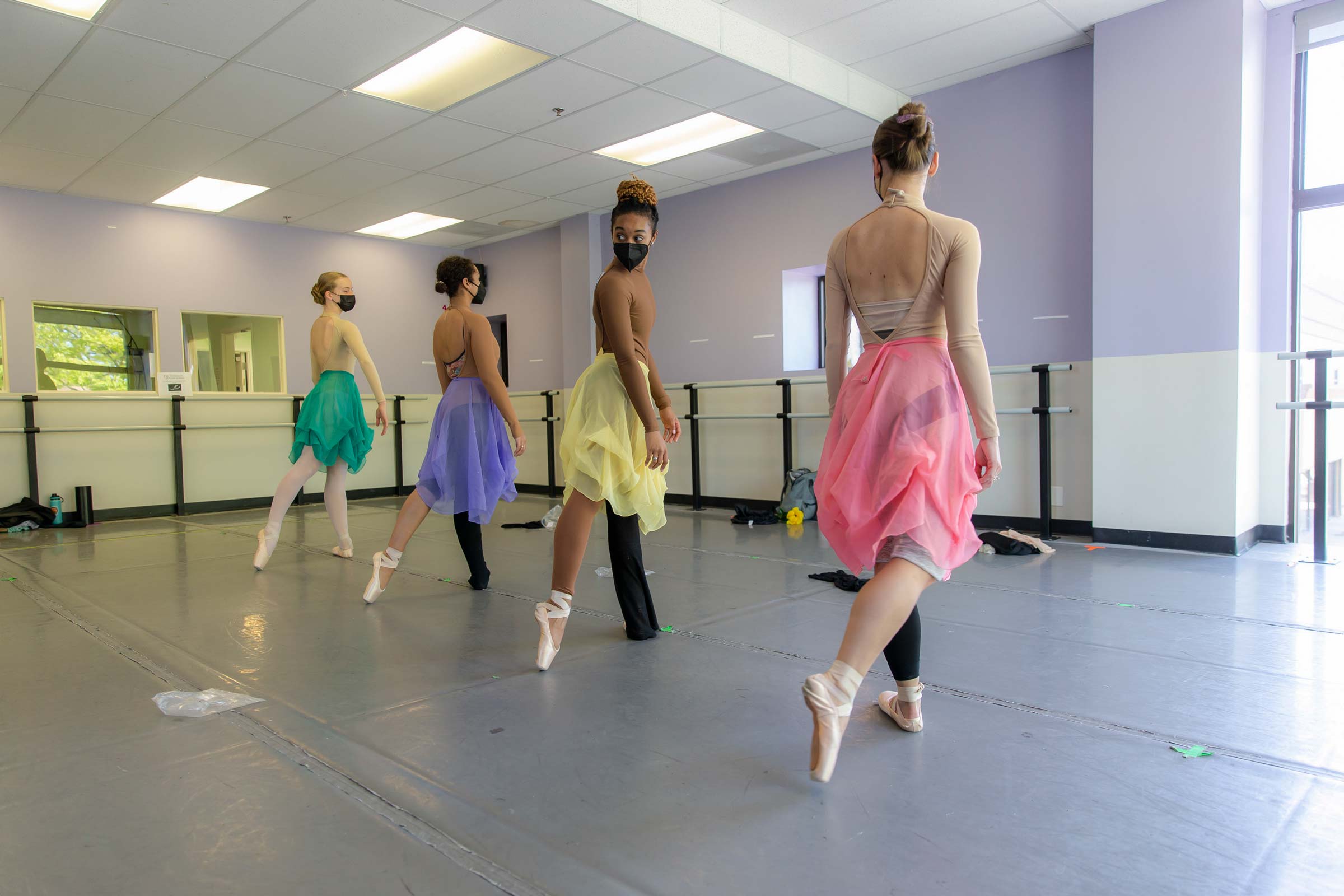 Four ballerinas in different colored skirts stand in a line, back toes pointed