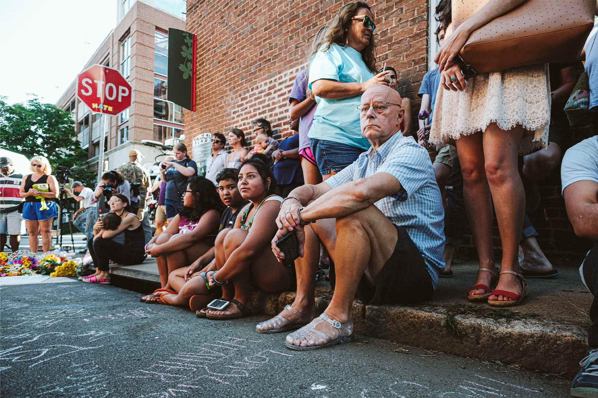 People sit on a curb in front of a brick building looking mournful.