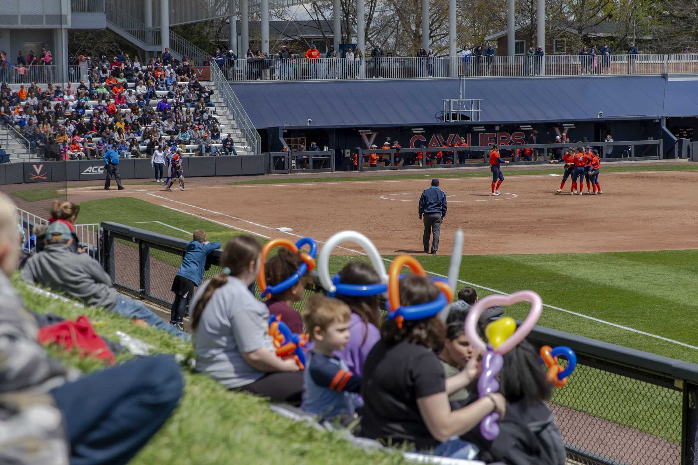 A group of children in balloon hats sit on a grassy hill, watching the softball game.
