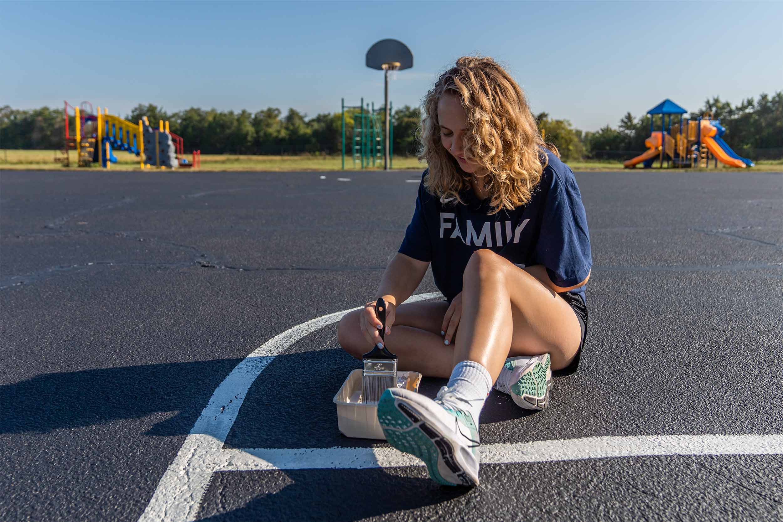 A volunteer uses a paint brush to repaint lines on an outdoor basketball court.