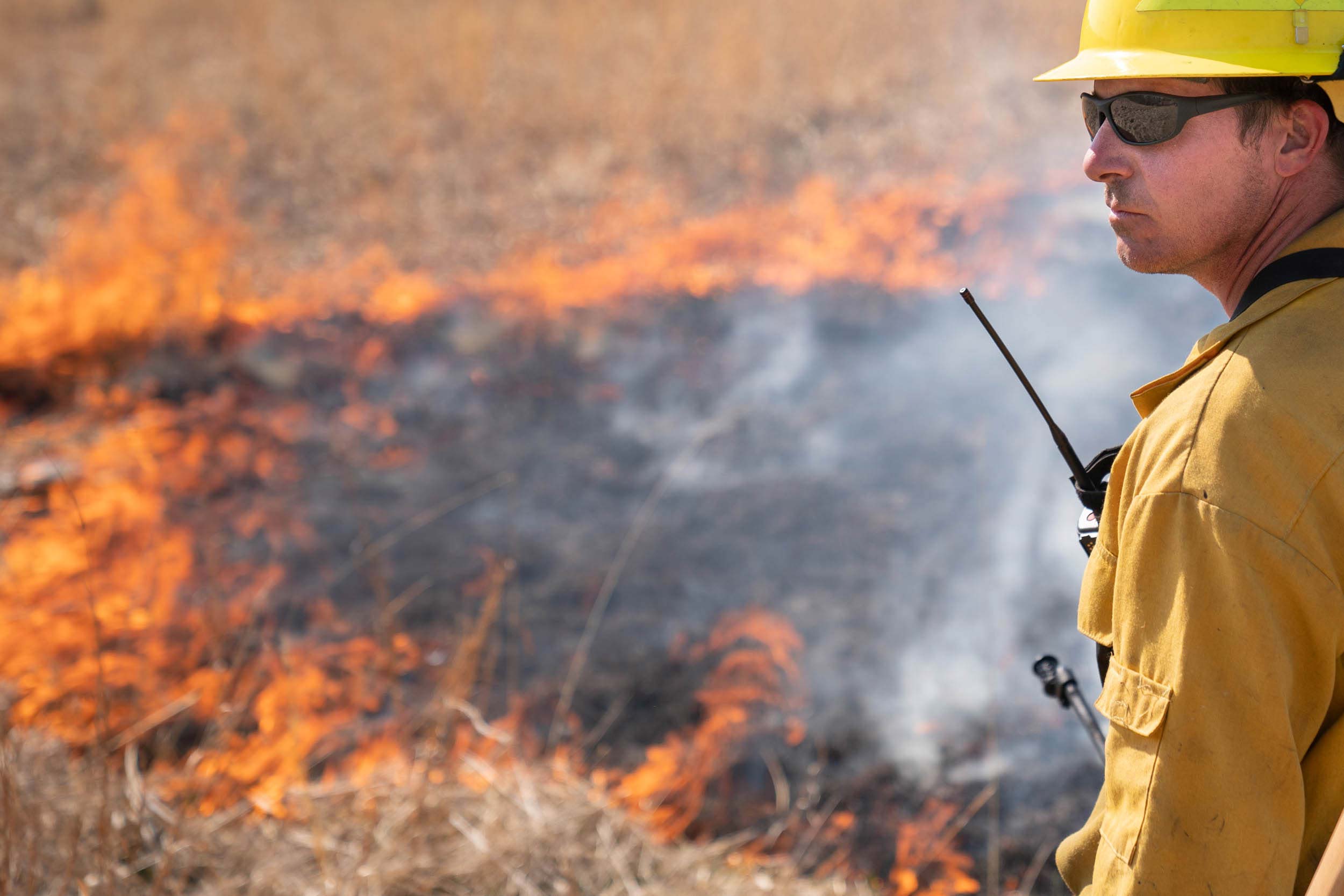 A man in a hard hat holds a walkie-talkie near a patch of burning thatch