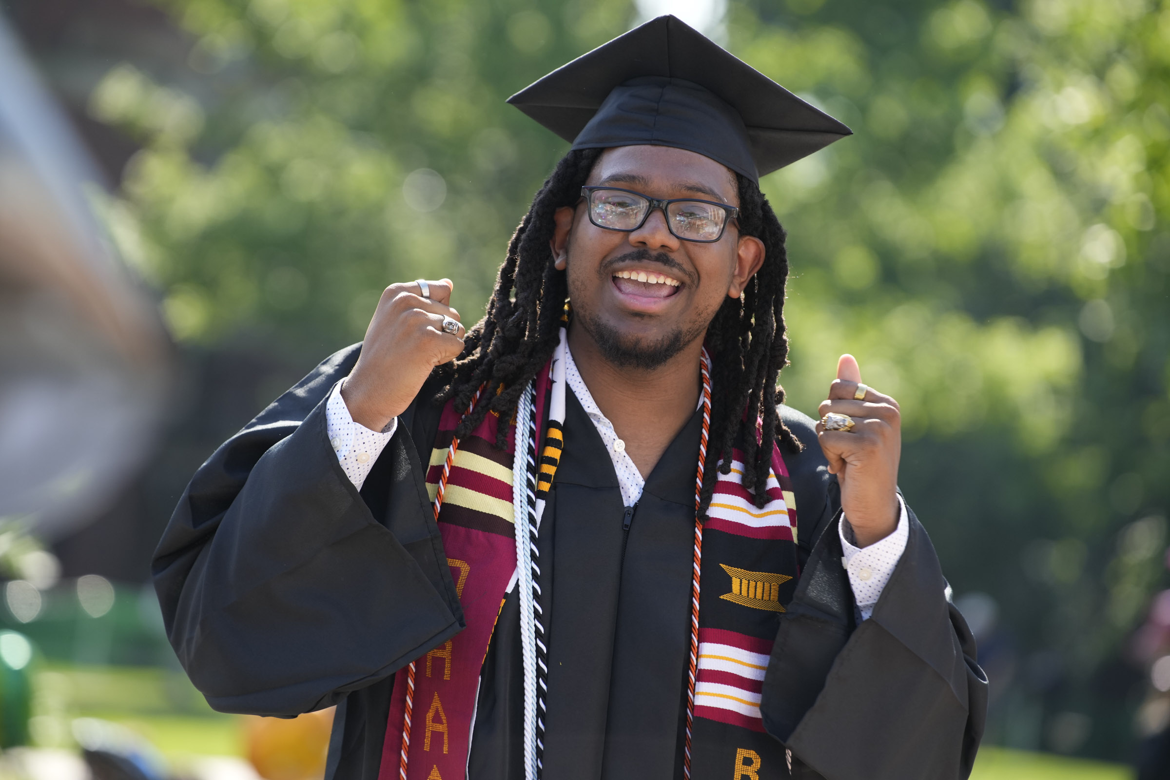 A student wearing a colorful sash and a graduation cap smiles a raises his hands. 